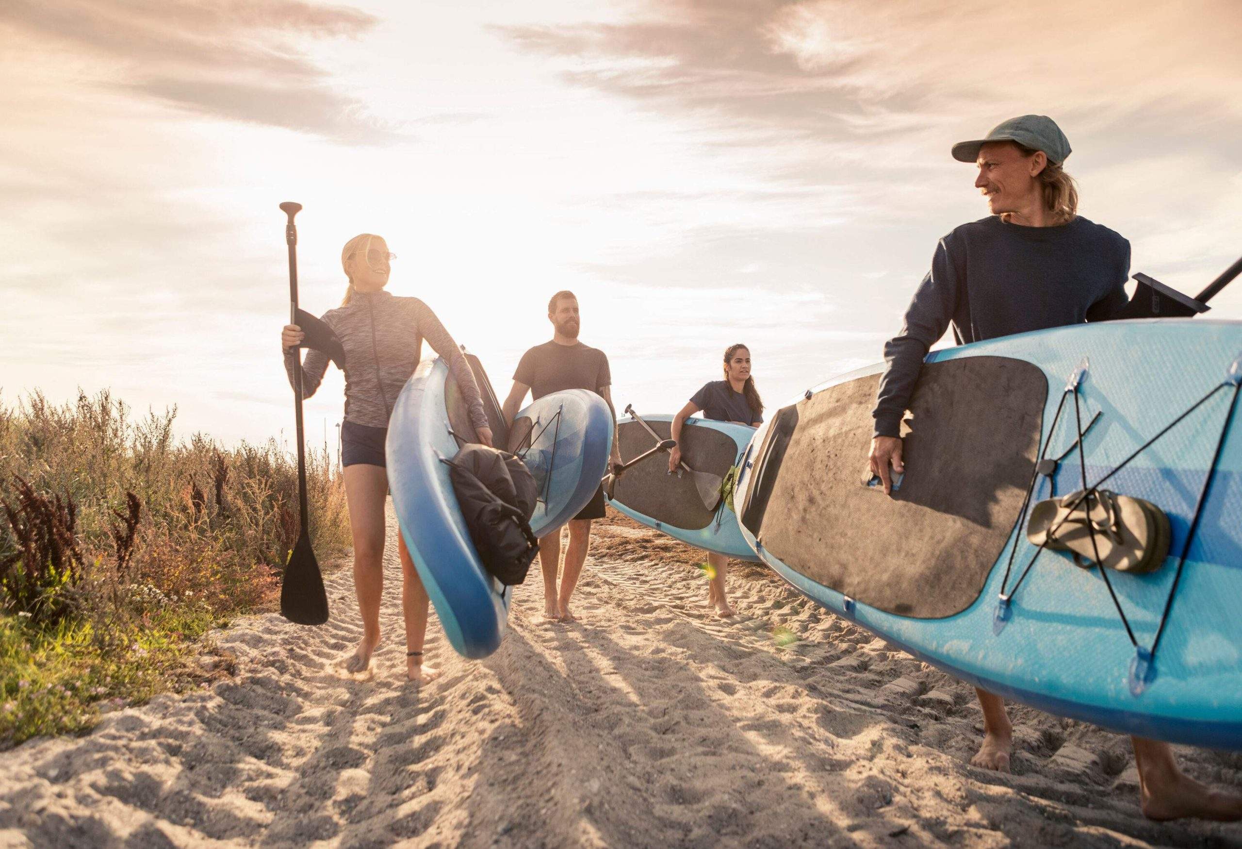 A group of four friends carries their large boards and paddles, walking along bushy sand on a sunny day.
