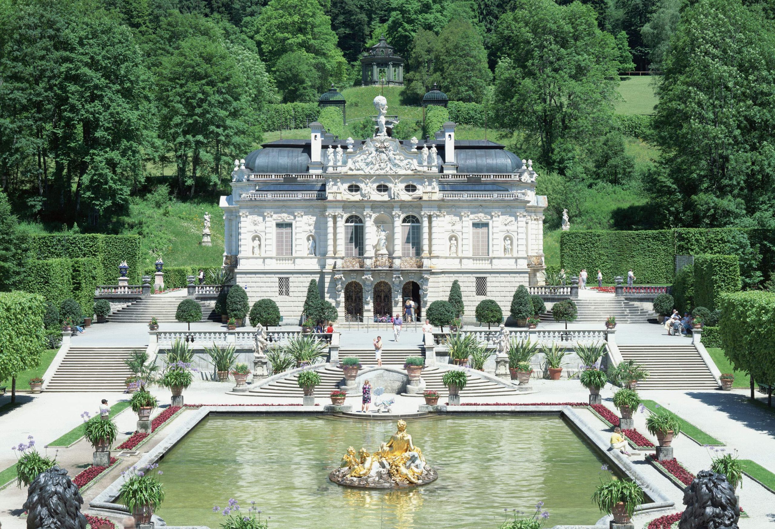 Linderhof Palace is a white palace with intricate stone façade and a grand front staircase in front of a pond with gold-plated statue in the centre.