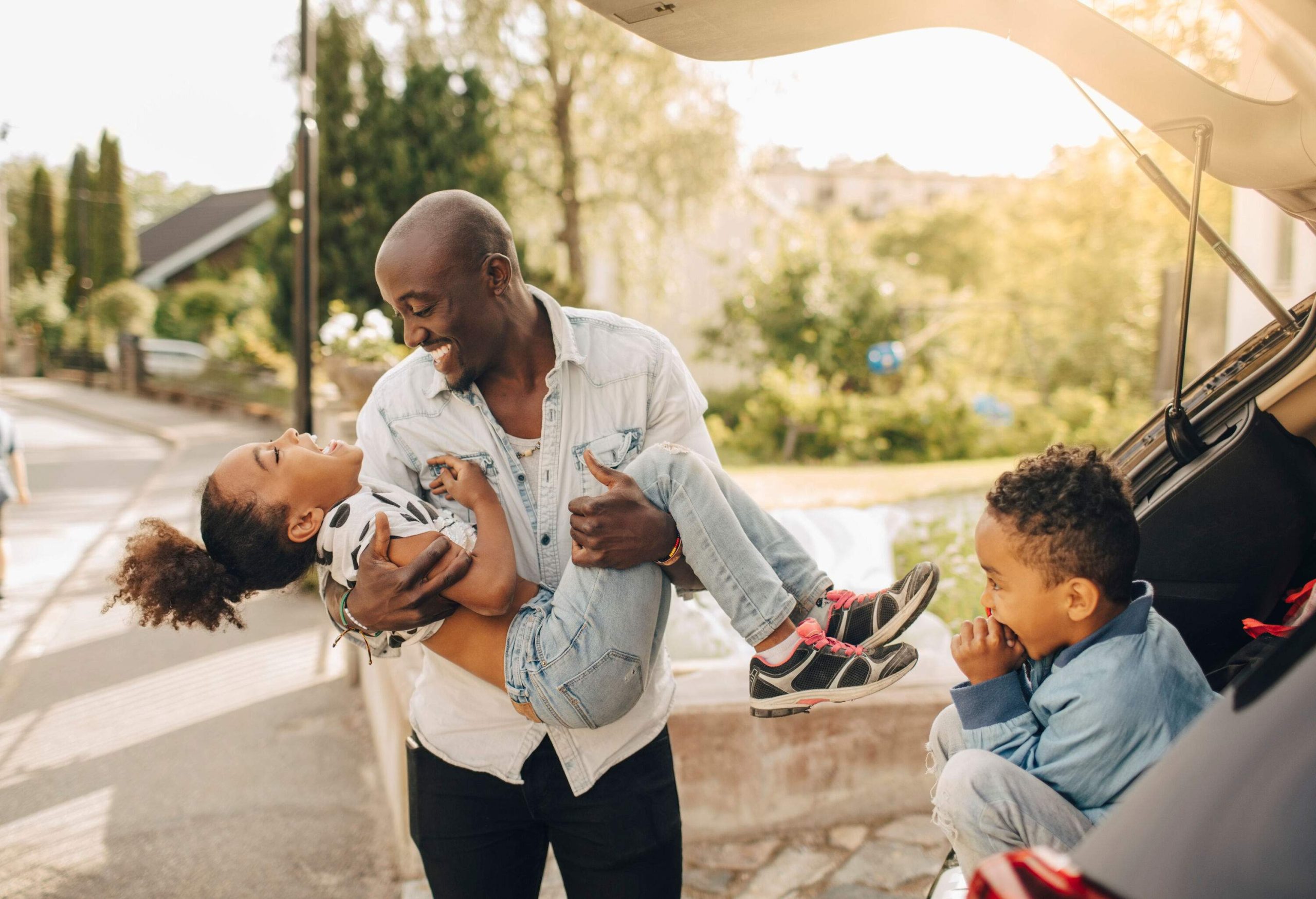 A man smiles as he carries a laughing child beside a young boy sitting on the trunk of a car.