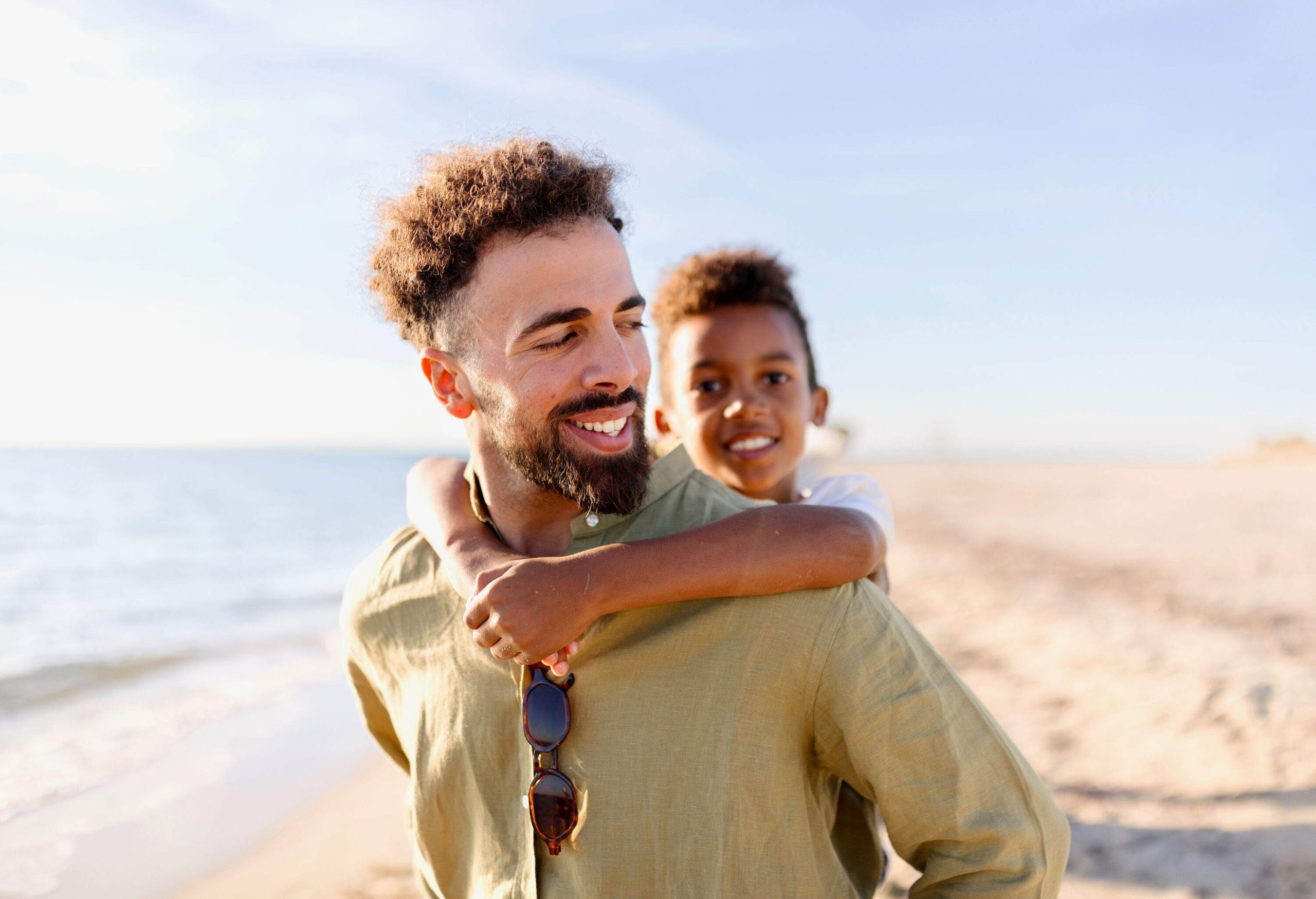 A man carrying a little boy on his back while walking on the beach.