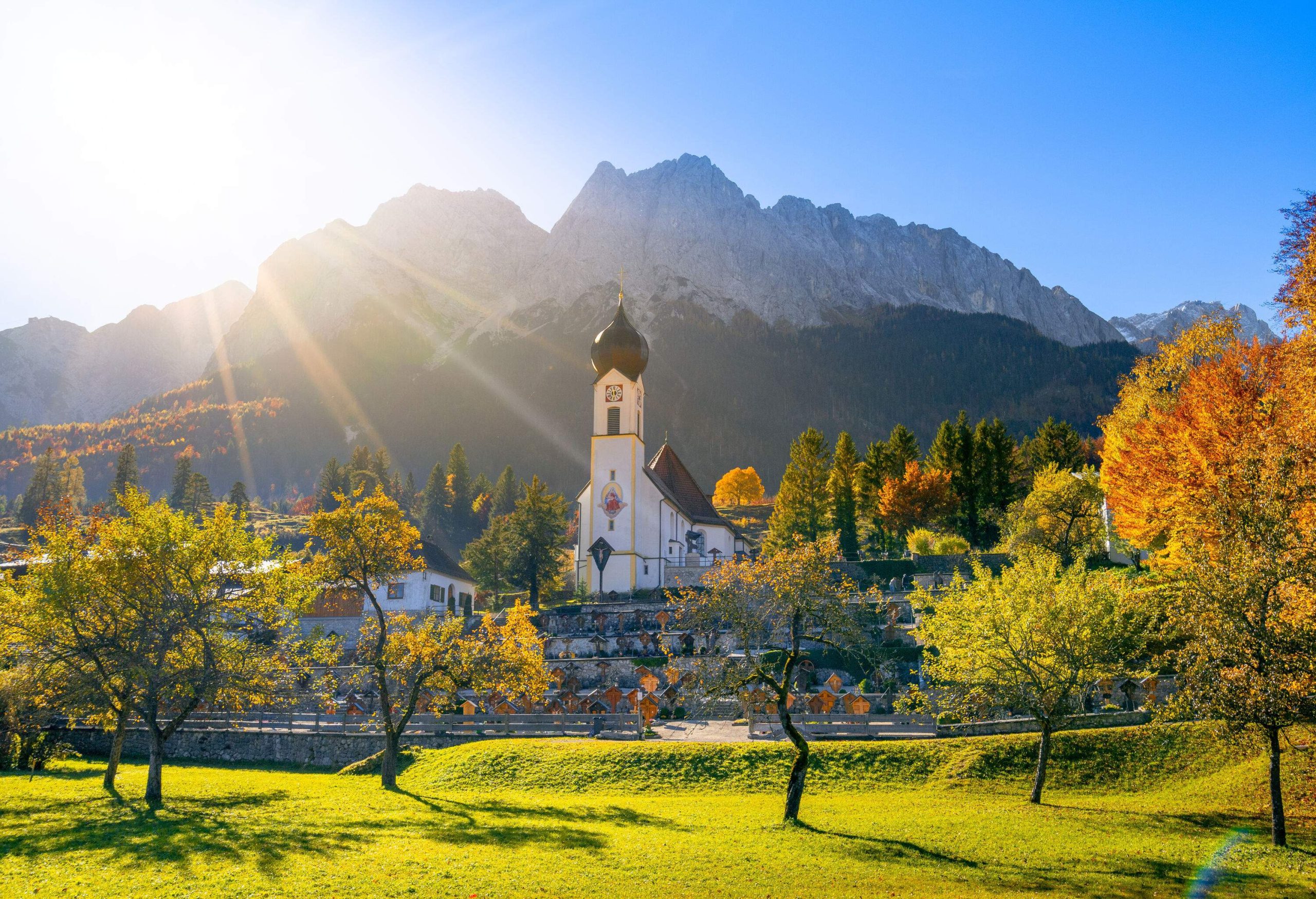 A white church with onion-shaped roof on a hill overlooking a green lawn with autumnal trees.