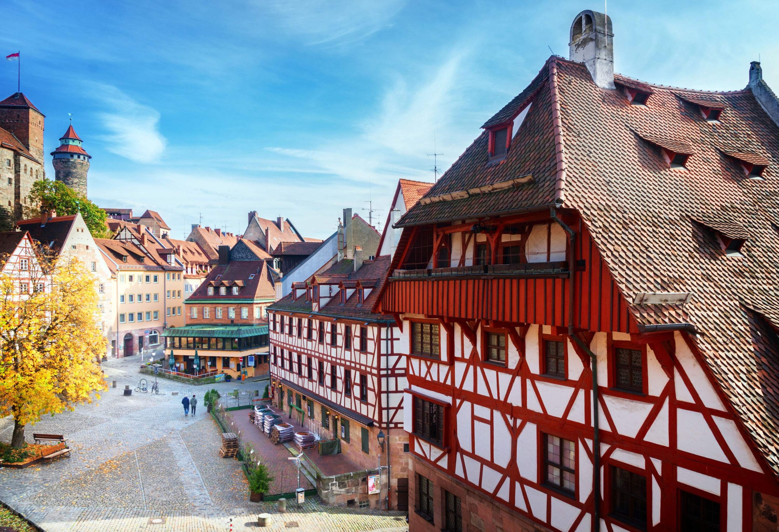 Two men walk on the cobbled street alongside lined tall colourful half-timbered buildings.