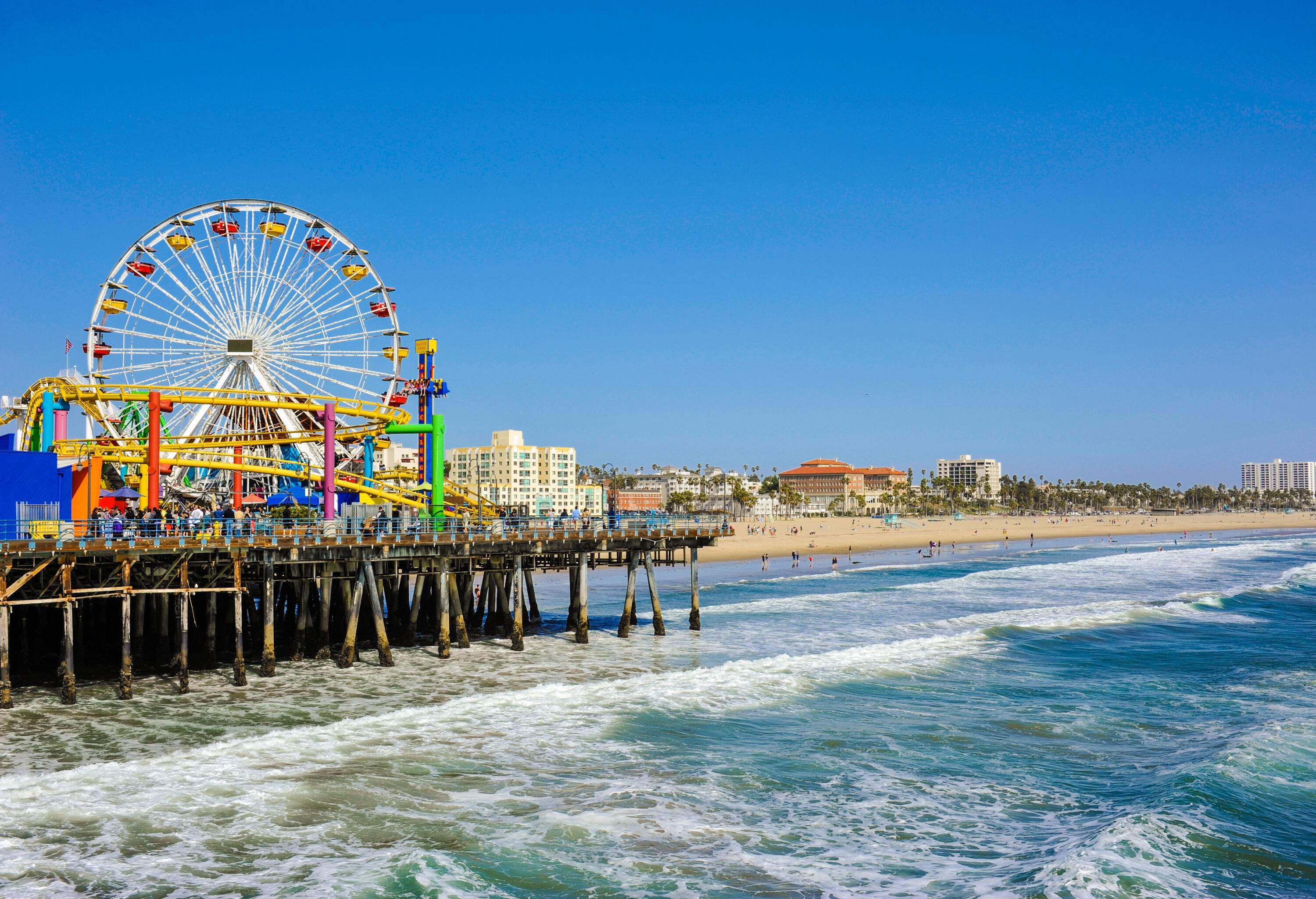 A pier with a colourful amusement park on the shore of a beach along an urban cityscape.
