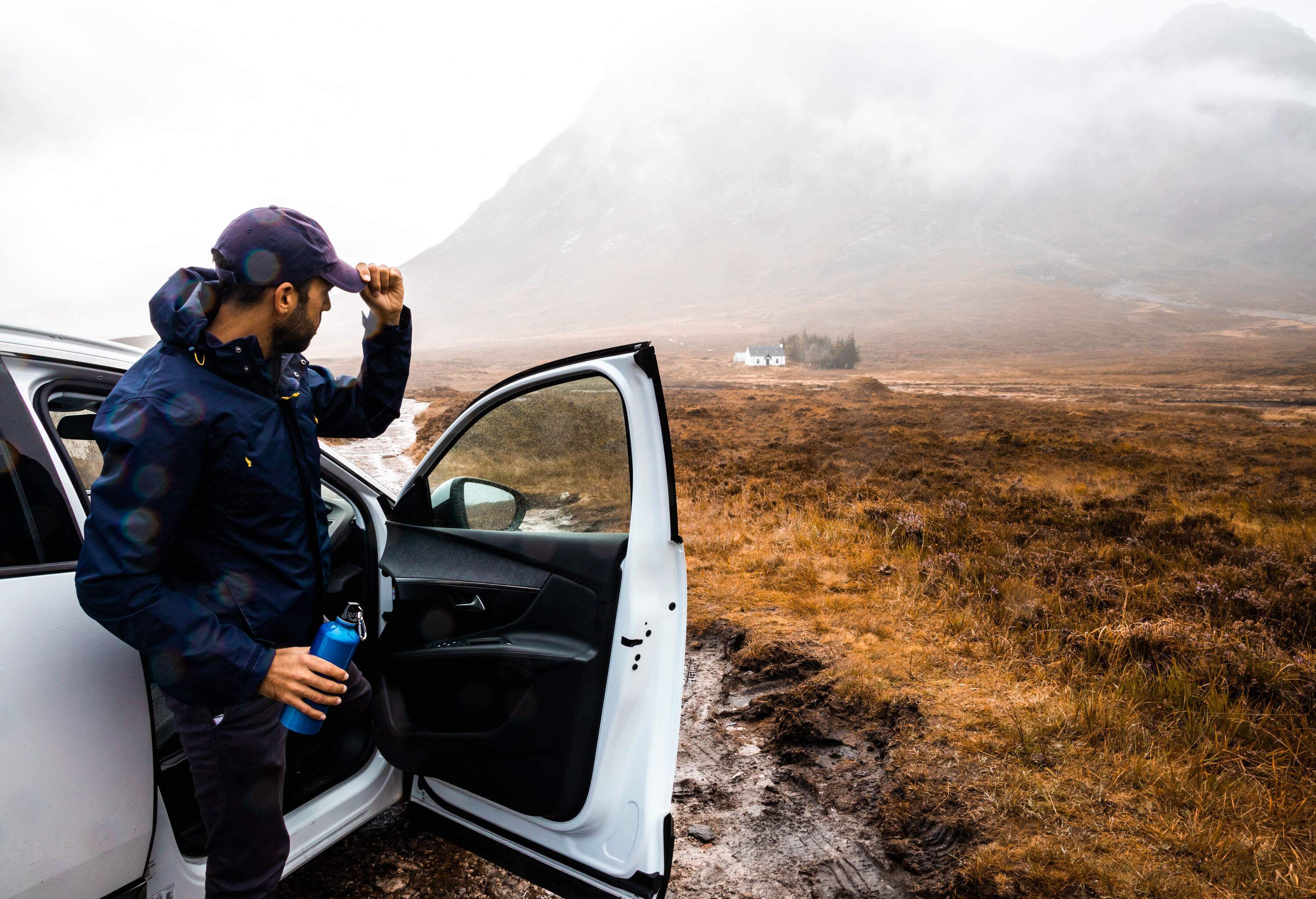 Man clad in a blue windbreaker emerges from a white car, holding a flask and looking at the bare landscape on a cloudy day