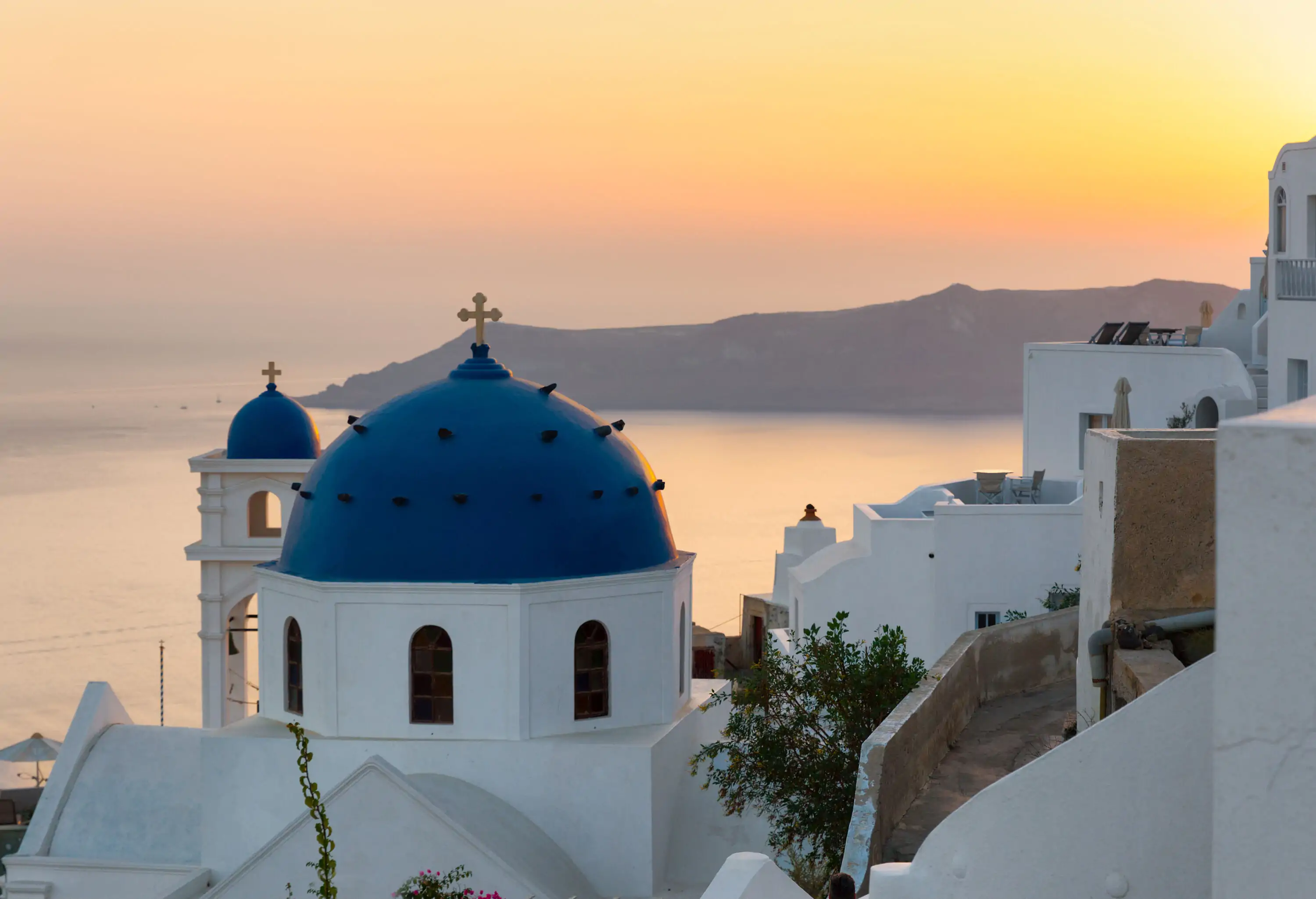 A white church with a blue dome roof and cross finial overlooks a vast ocean under an orange sky.
