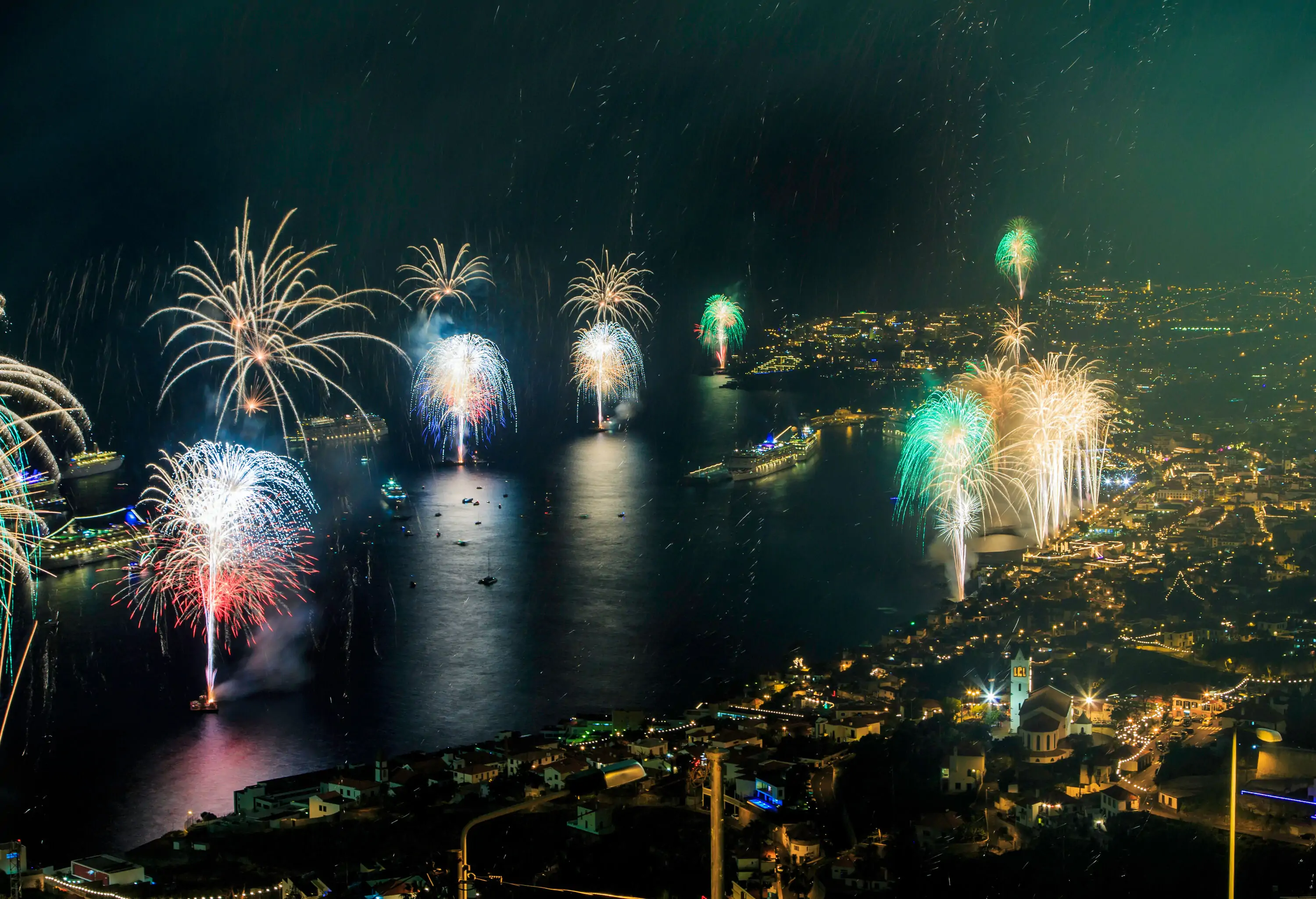 Colourful fireworks over the sea and the brightly lit coastal city against the night sky.