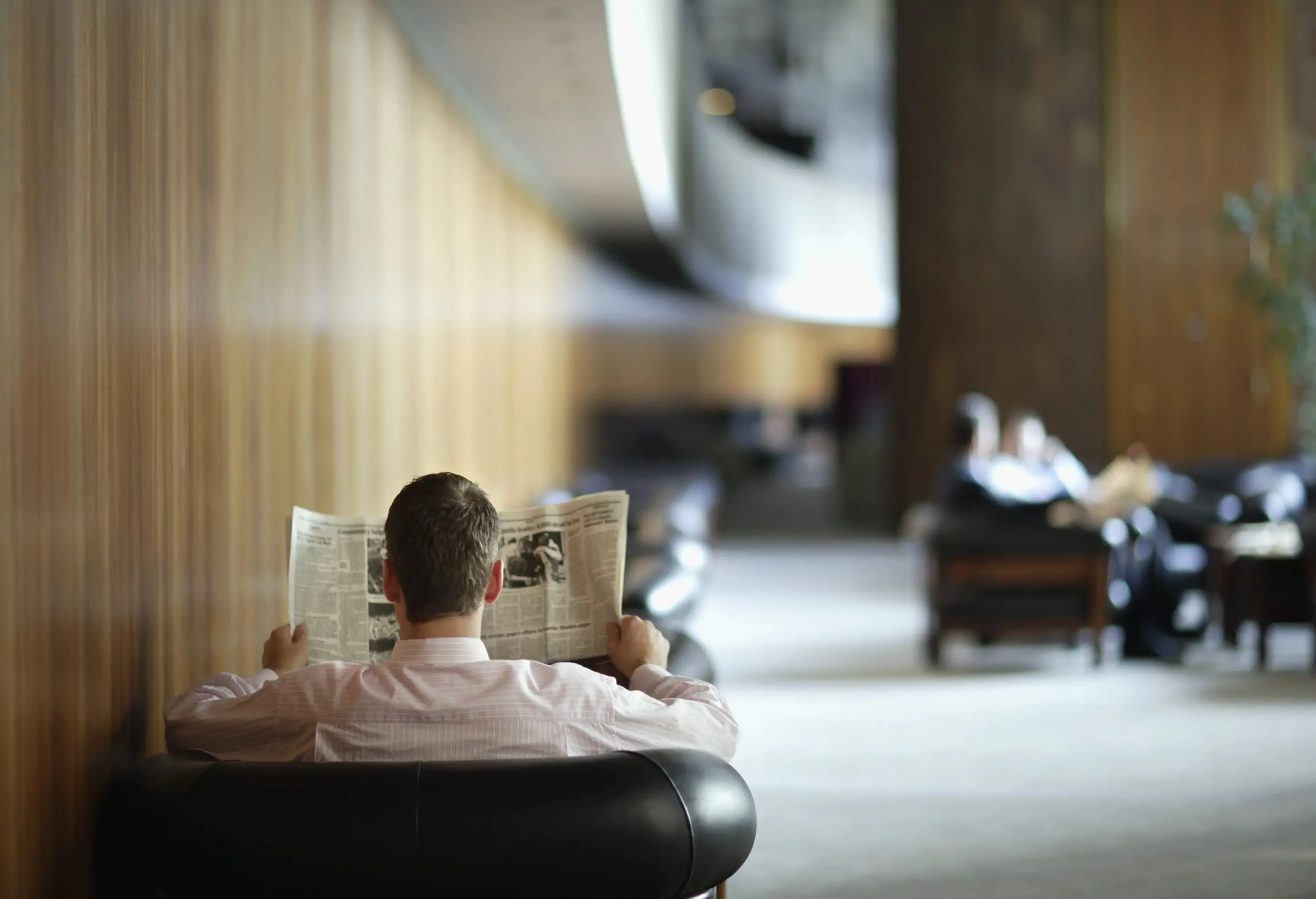 A man sits comfortably in a hotel lobby, reading a newspaper. The lobby features modern decor with a cozy seating area, warm lighting, and a calm ambiance.