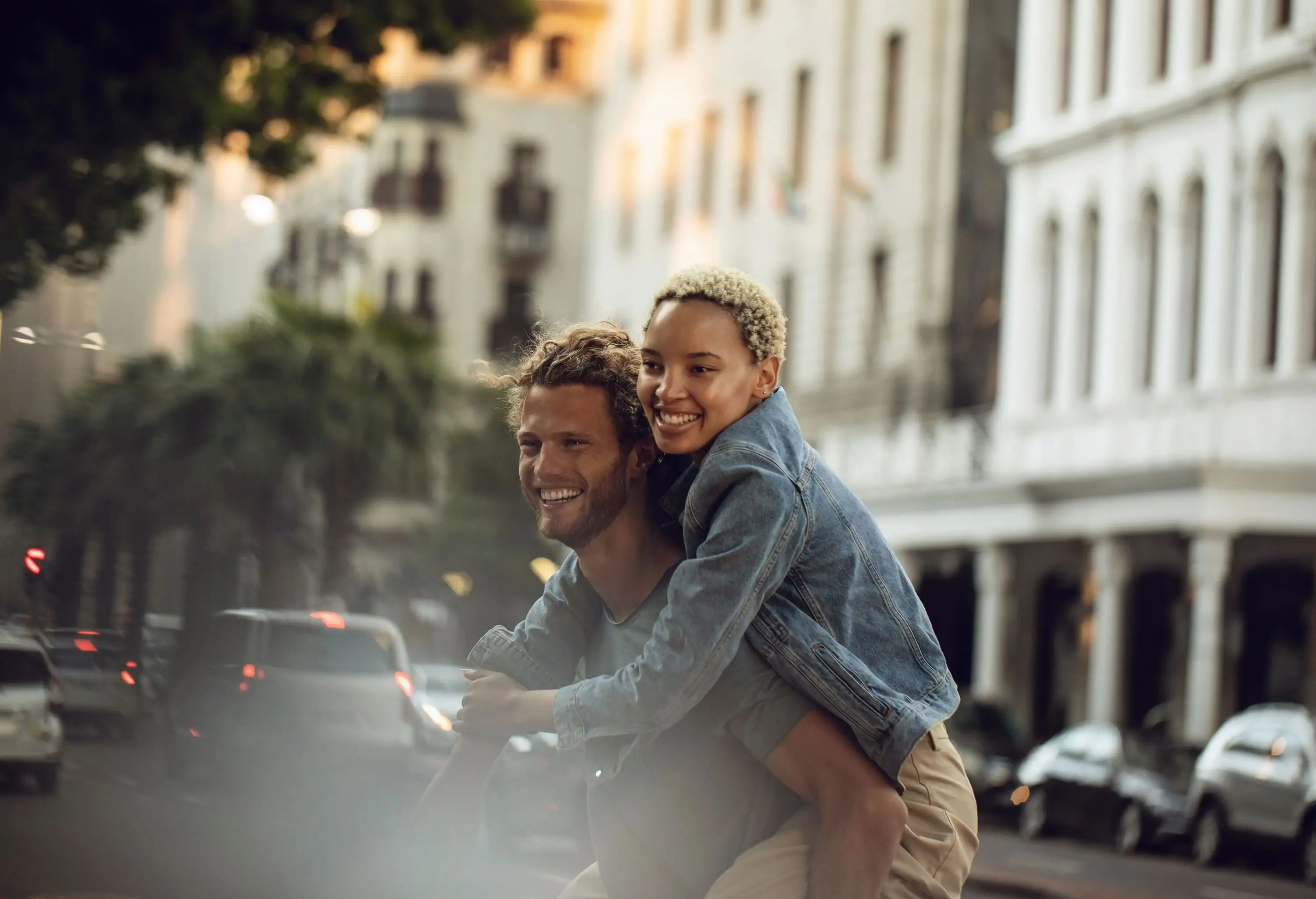 A man gives a piggyback ride to his girlfriend while crossing a street alongside tall buildings.