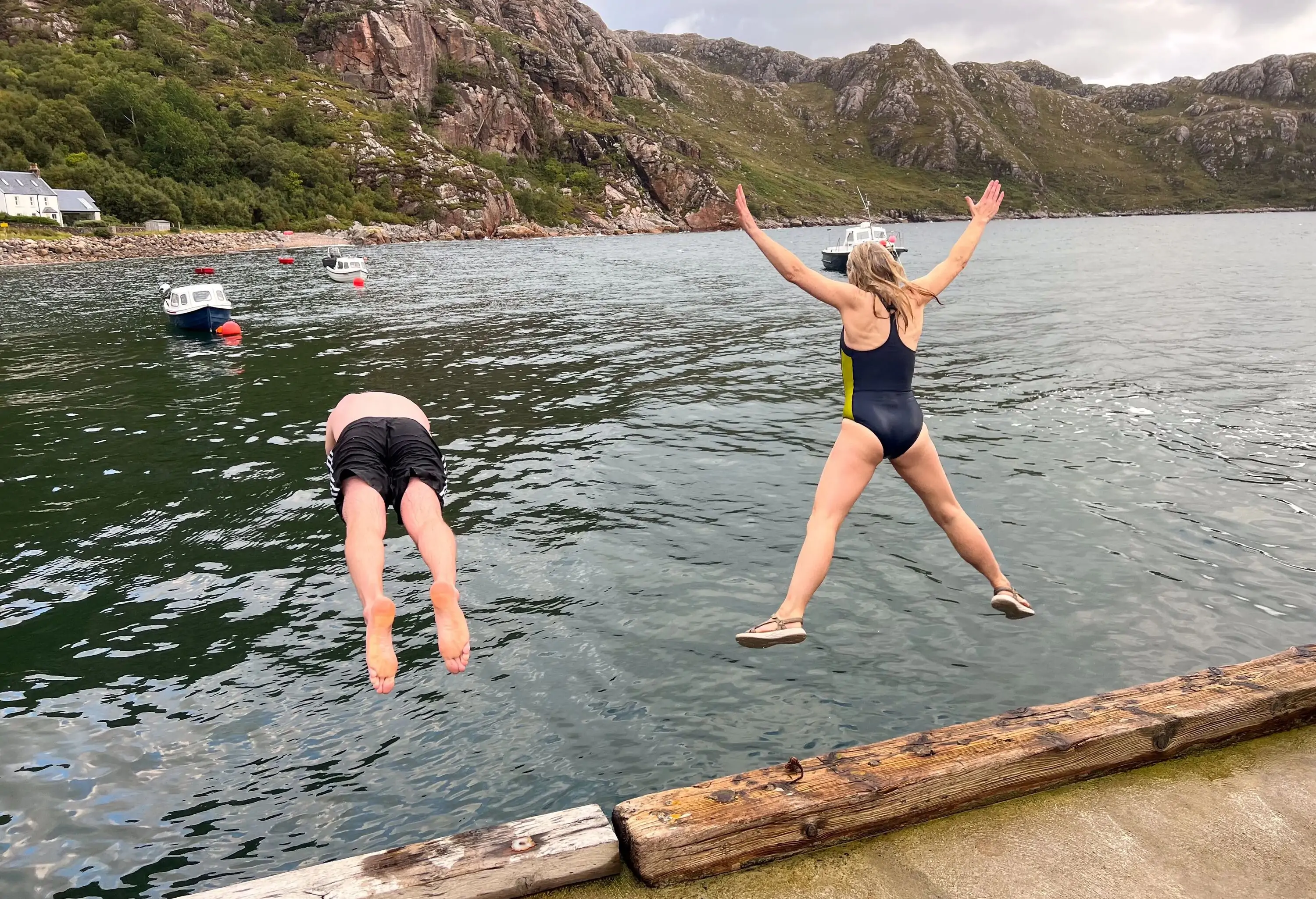 A full shot of couple mid-air jumping into a loch at the same time on the side of Loch Torridon near the village of Diabaig in the west highlands of Scotland. They are wearing swimming gear.