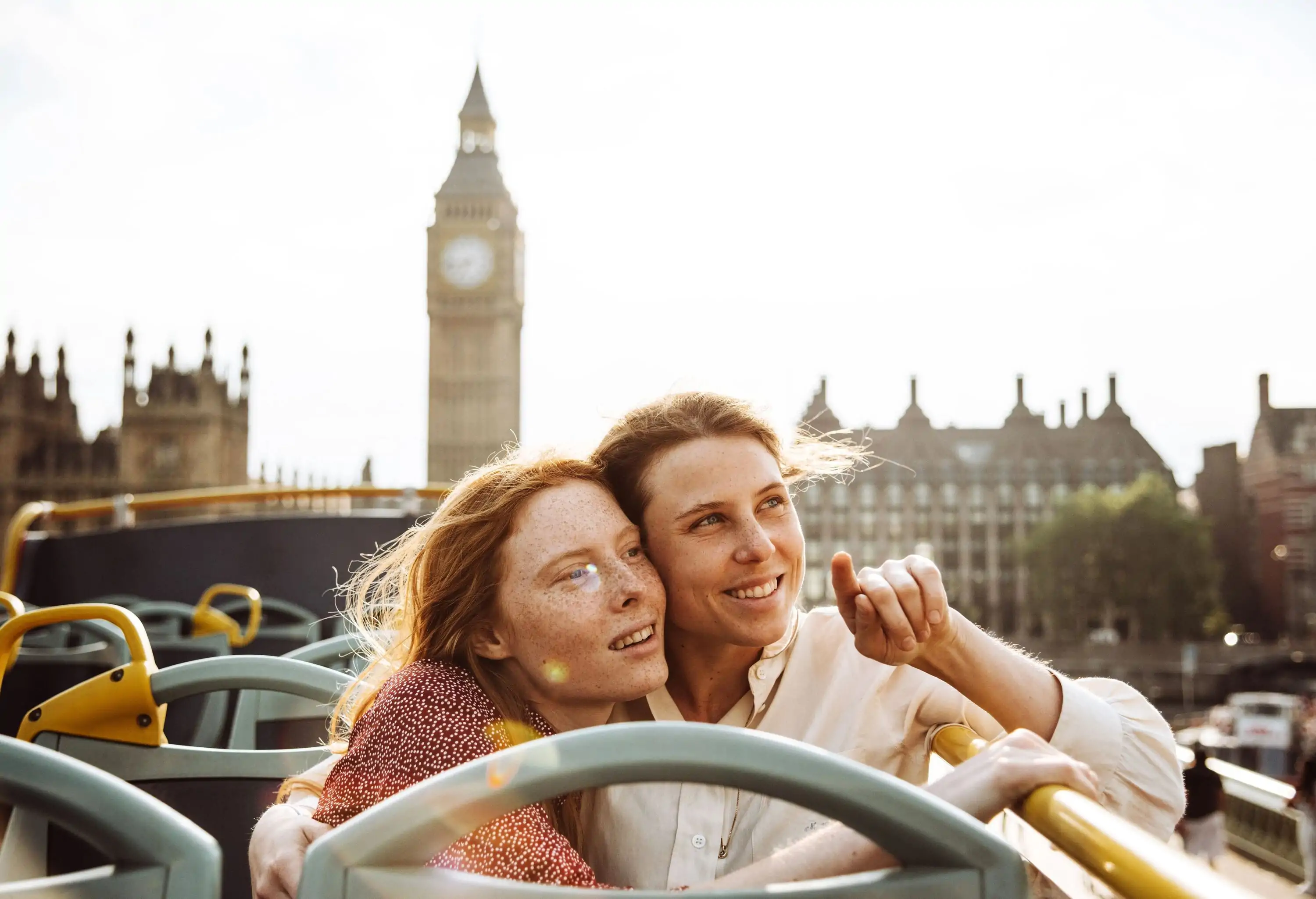 Female lovers sit and hug on an open-top bus while admiring the cityscape.