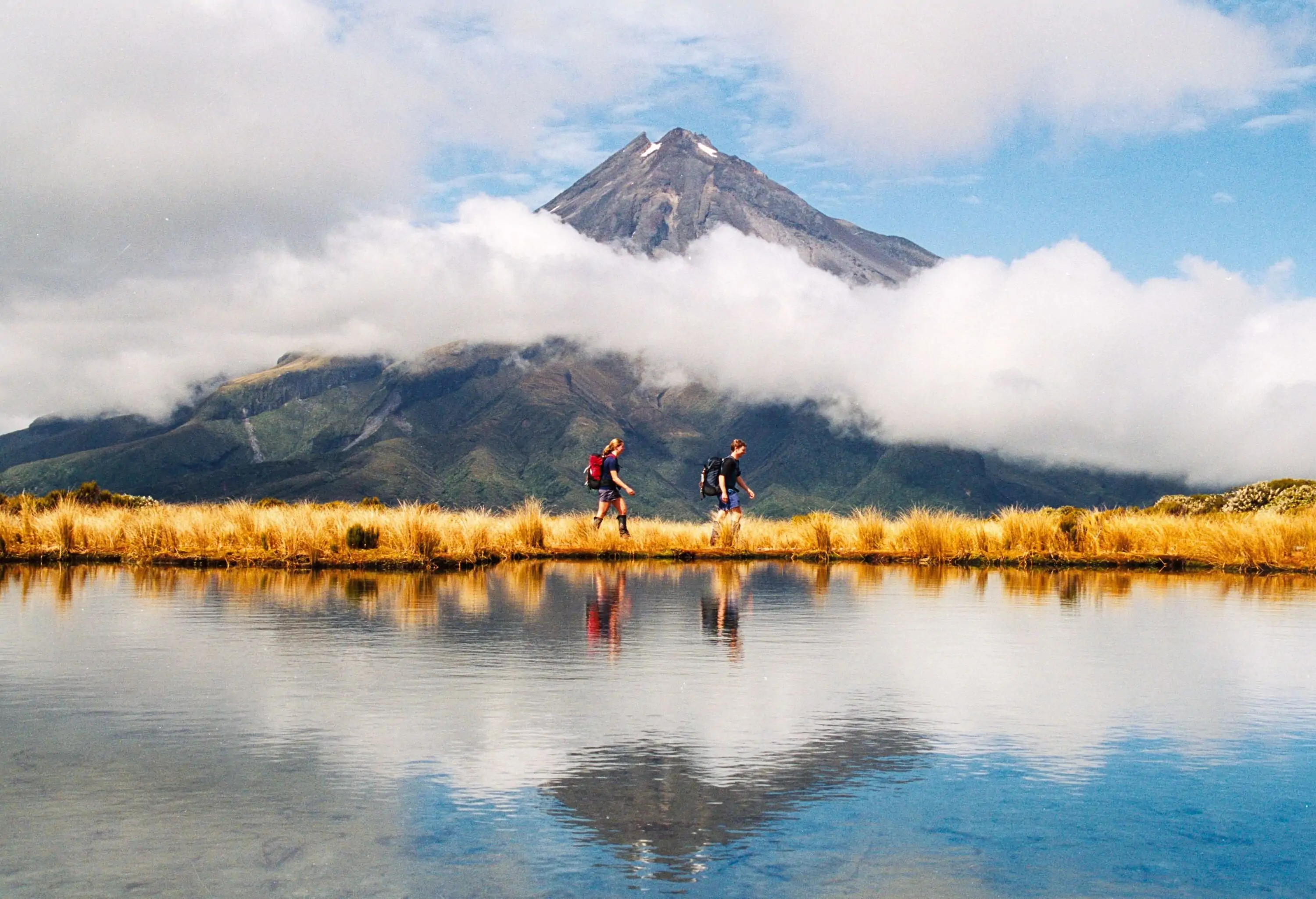 Two people hiking on a grassy area next to a lake with a cloud-covered mountain in the background.