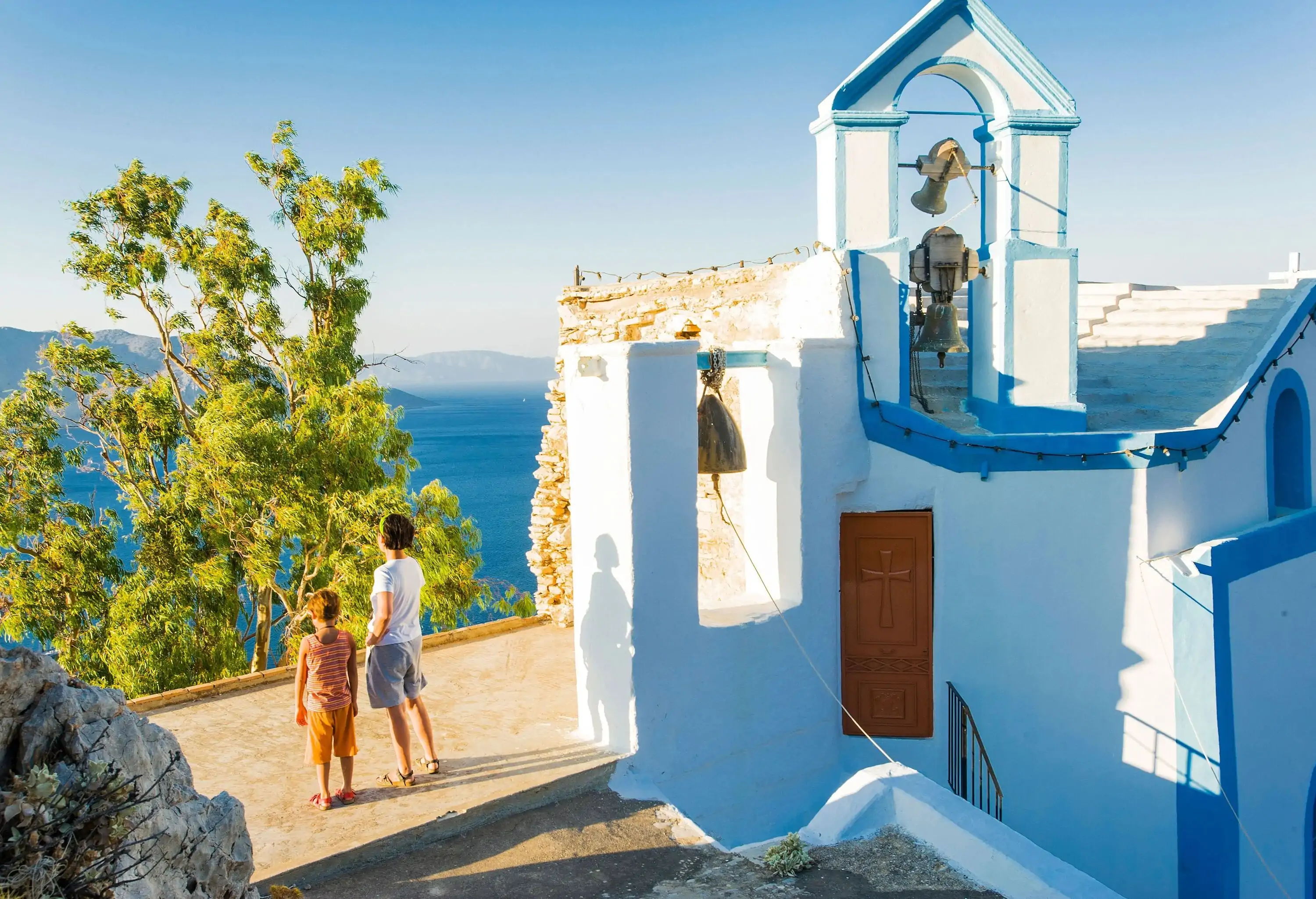 Two individuals stand on the side of a traditional white Greek chapel overlooking the blue sea.
