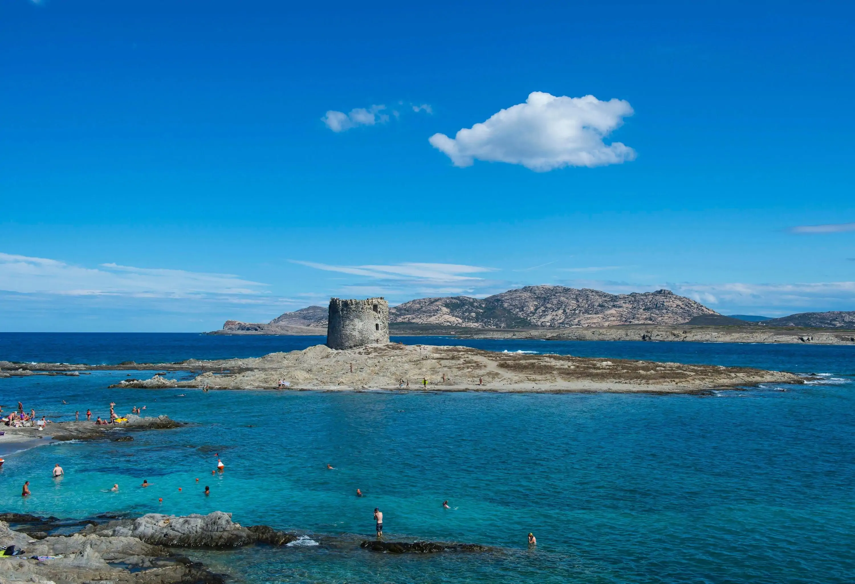 A medieval tower emerges on a rocky outcrop above the sea, and people swim on the surrounding turquoise water.