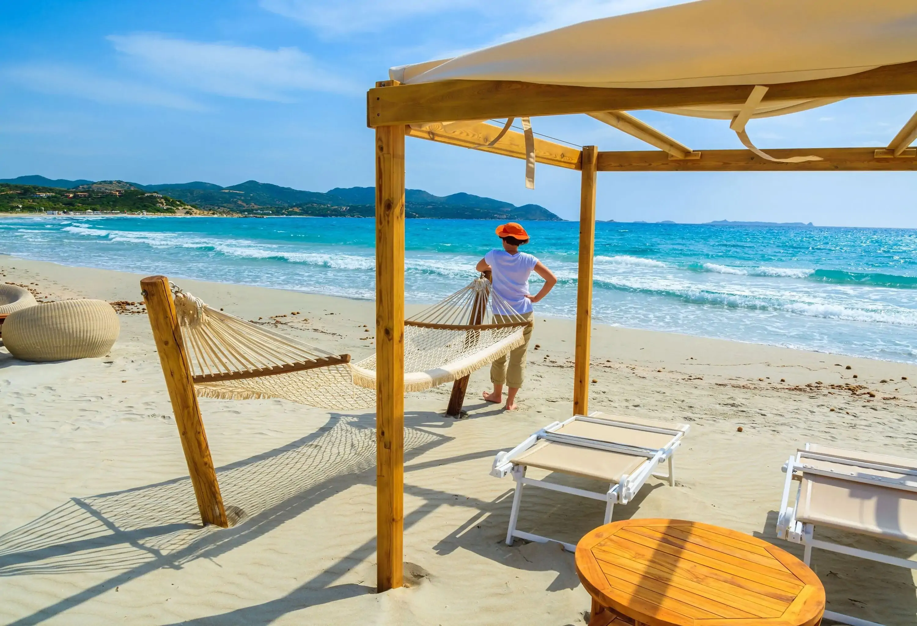 A woman leaning on a hammock stands along a shed with sunbeds and a wooden table on a relaxing white sand beach.