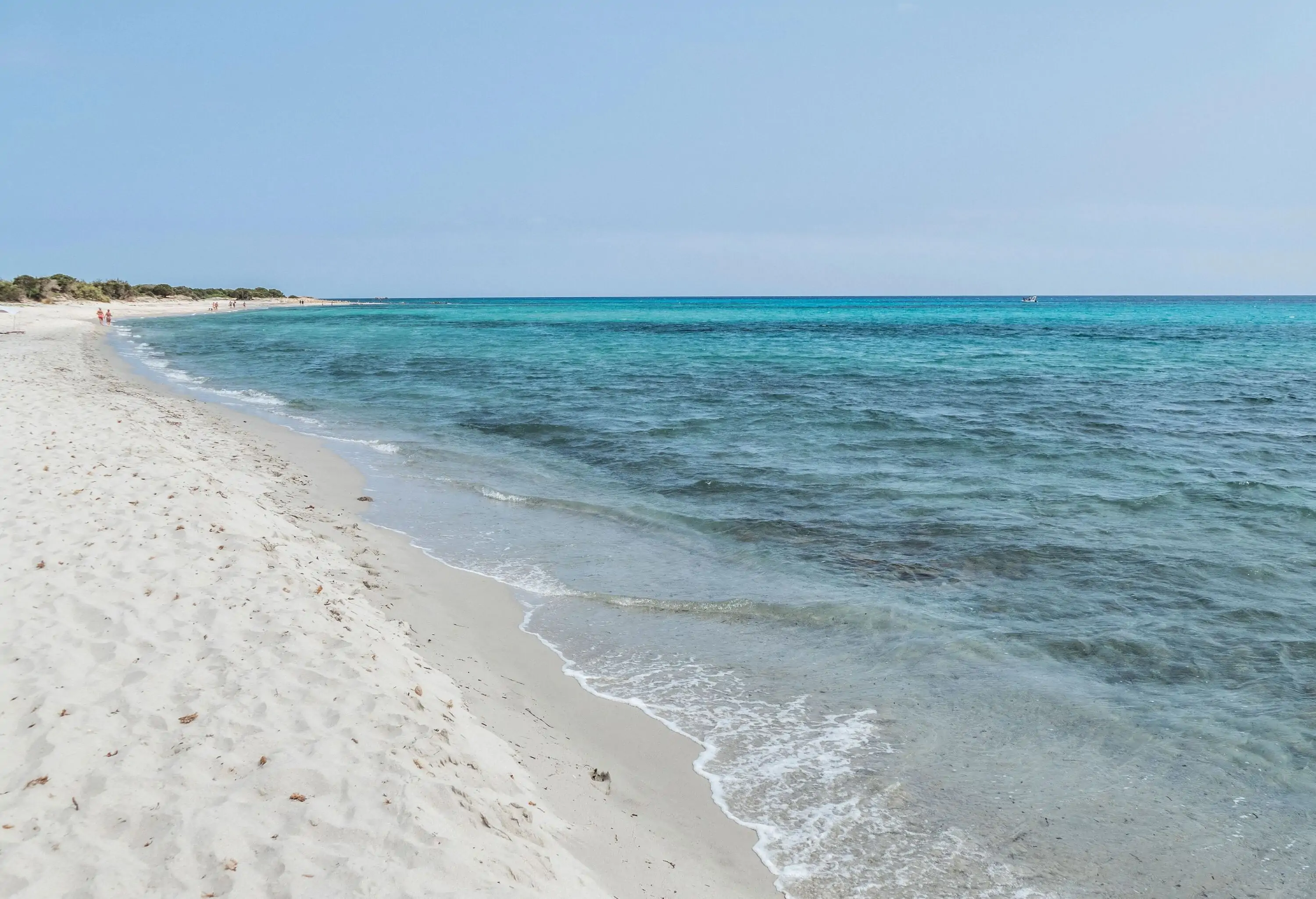 A long stretch of fine white sand beach with clear, calm turquoise water under a cloudless sky.