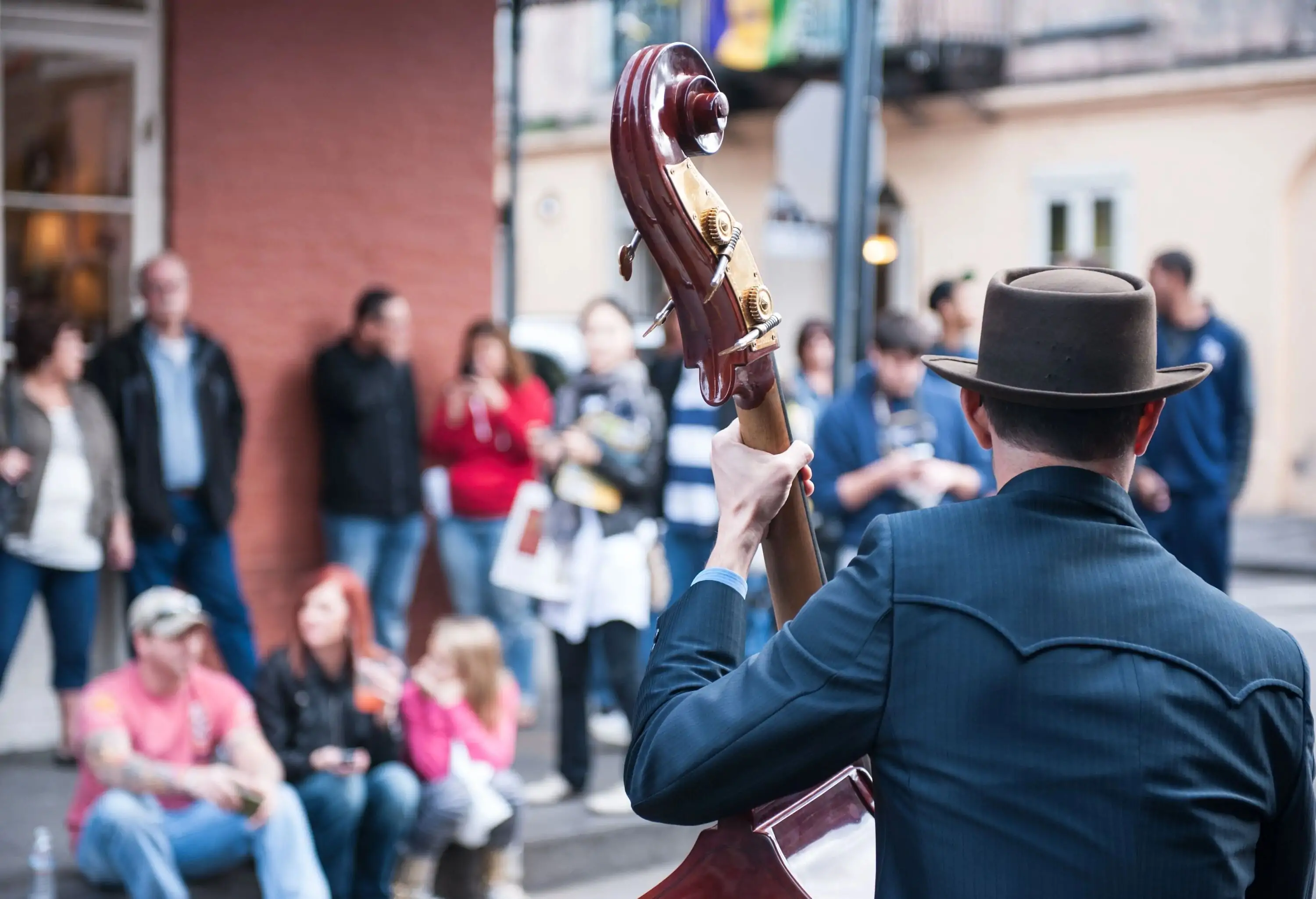 A man in a hat playing a string instrument while people on the sidewalk listen to its symphony.