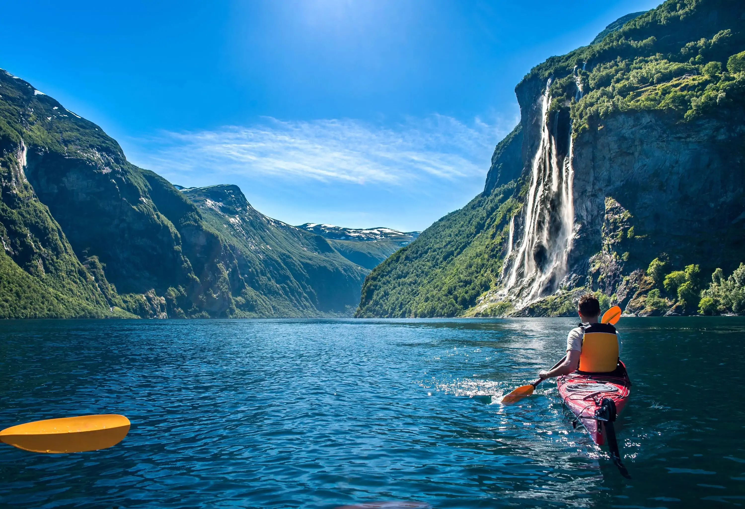 A man in a kayak paddling towards a waterfall on the mountainside on a tranquil lake surrounded by high mountains.