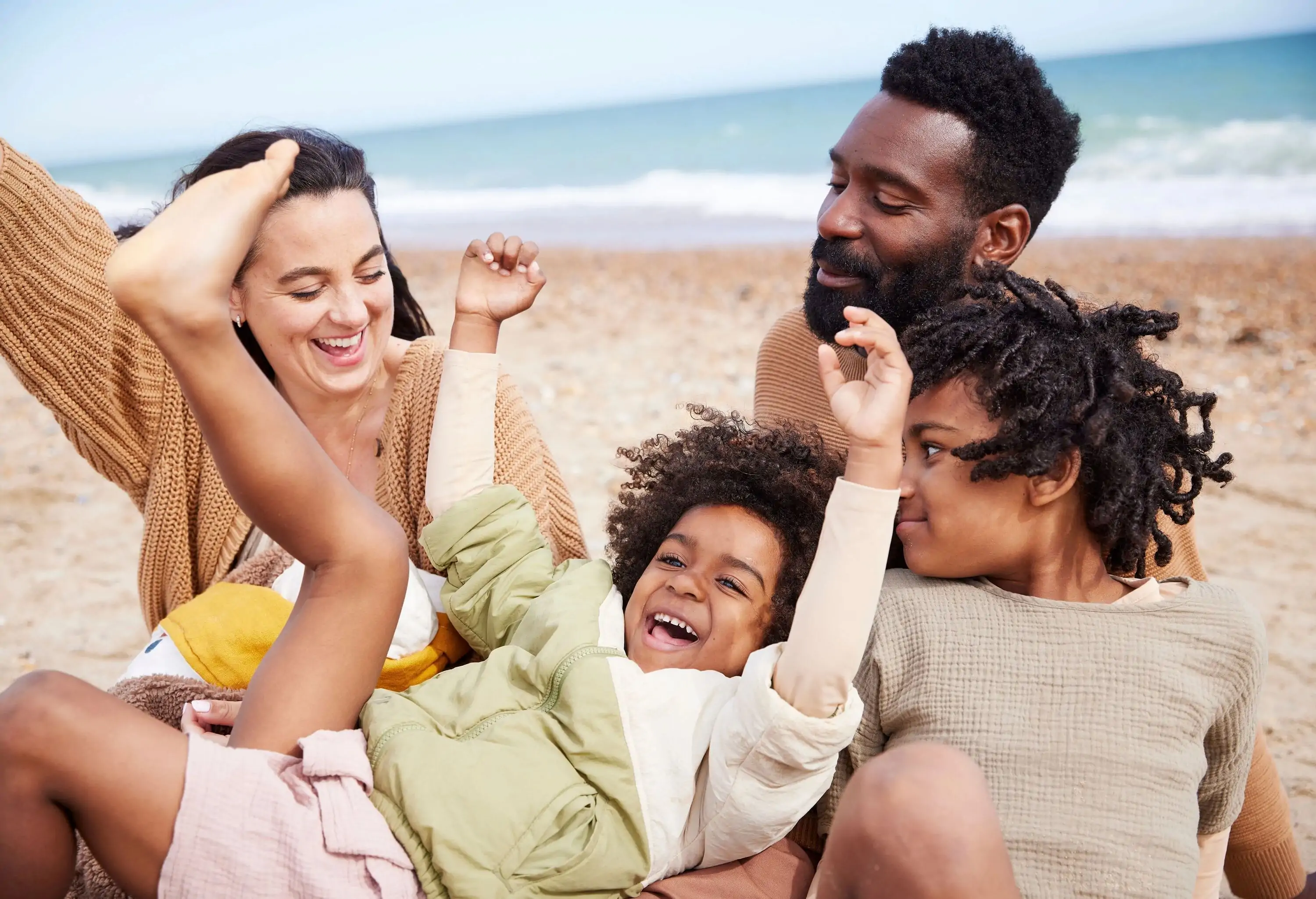 A family of 5 whooping with joy on the beach. A road trip to the coast, especially enjoyed by the youngest boy and mother. Arms raised and laughing, the youngest boy is laid back on the legs of his father. Oldest son looks amused by the scene.