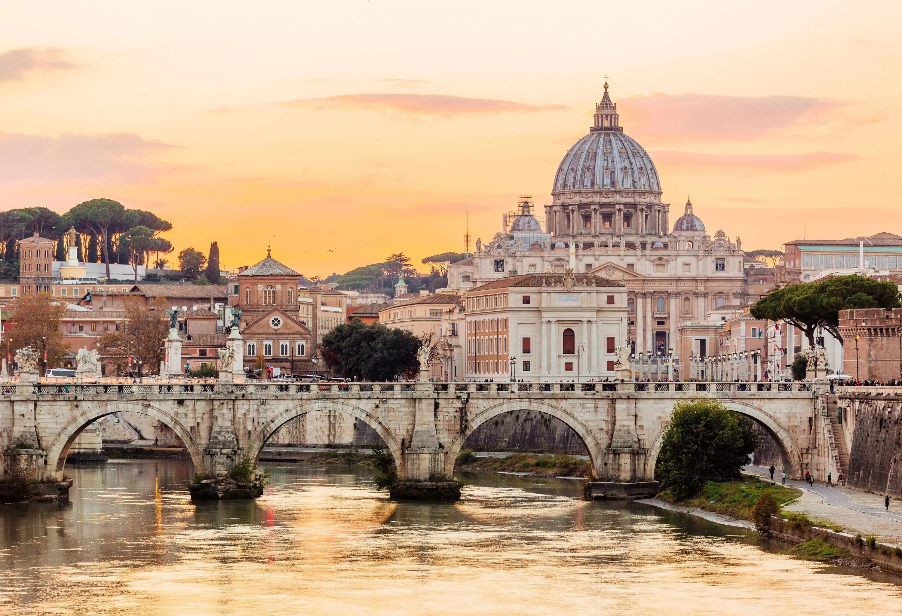 An arch bridge across a calm river with distant views of a domed church rising above a city under an orange sky.