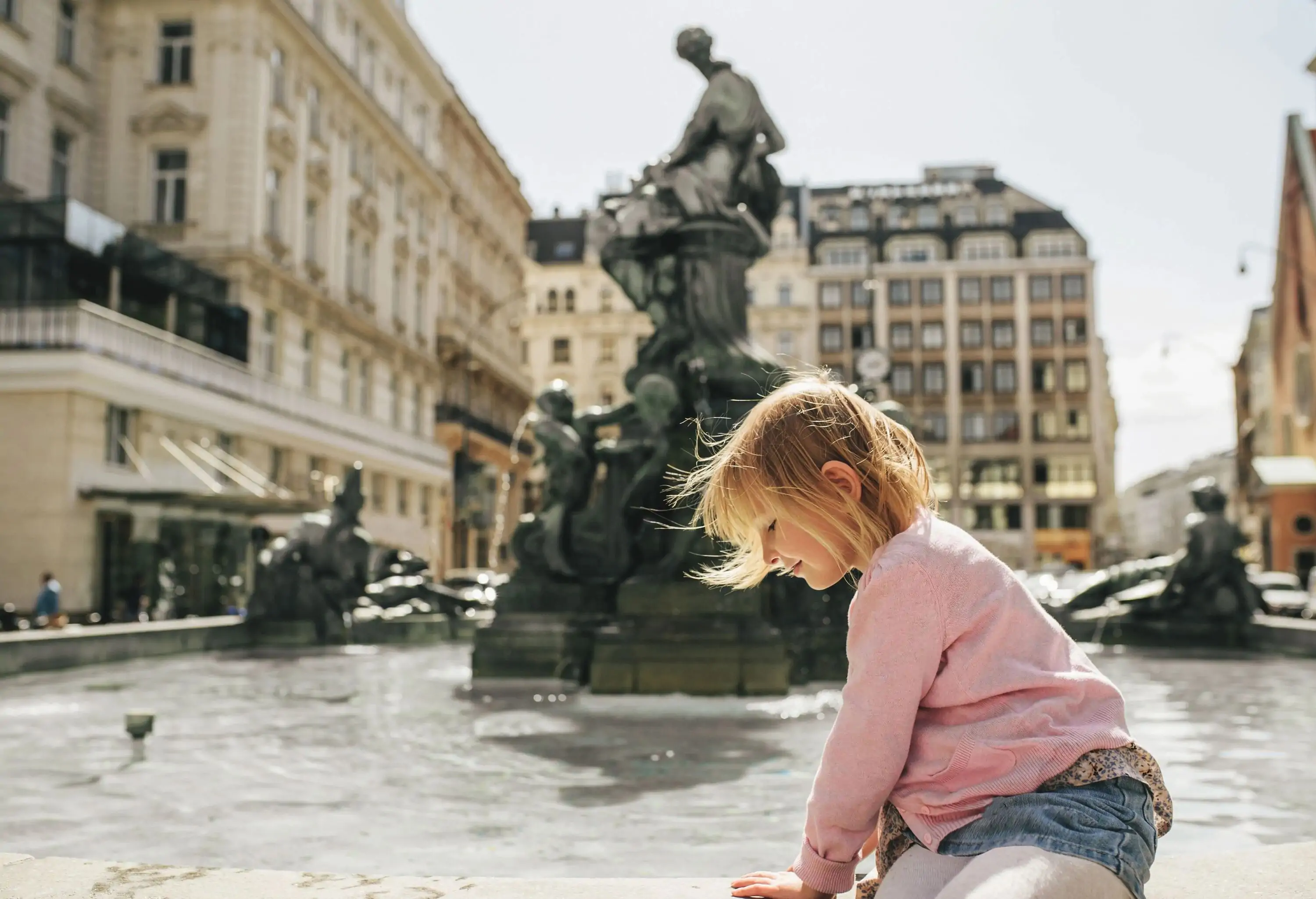 A little girl in a pink sweater playing in a water fountain.