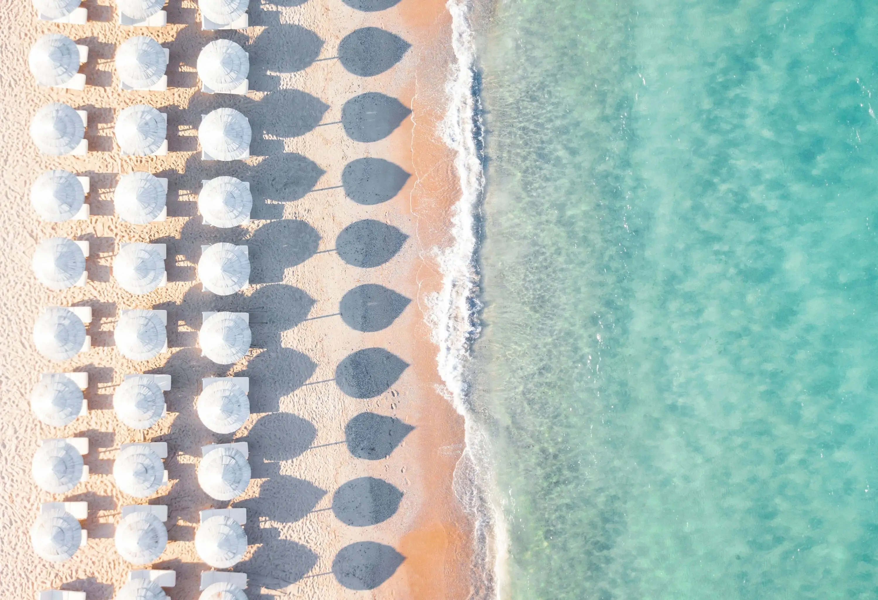 Rows of white umbrellas casting shadows on the shore where the turquoise sea meets the sand.