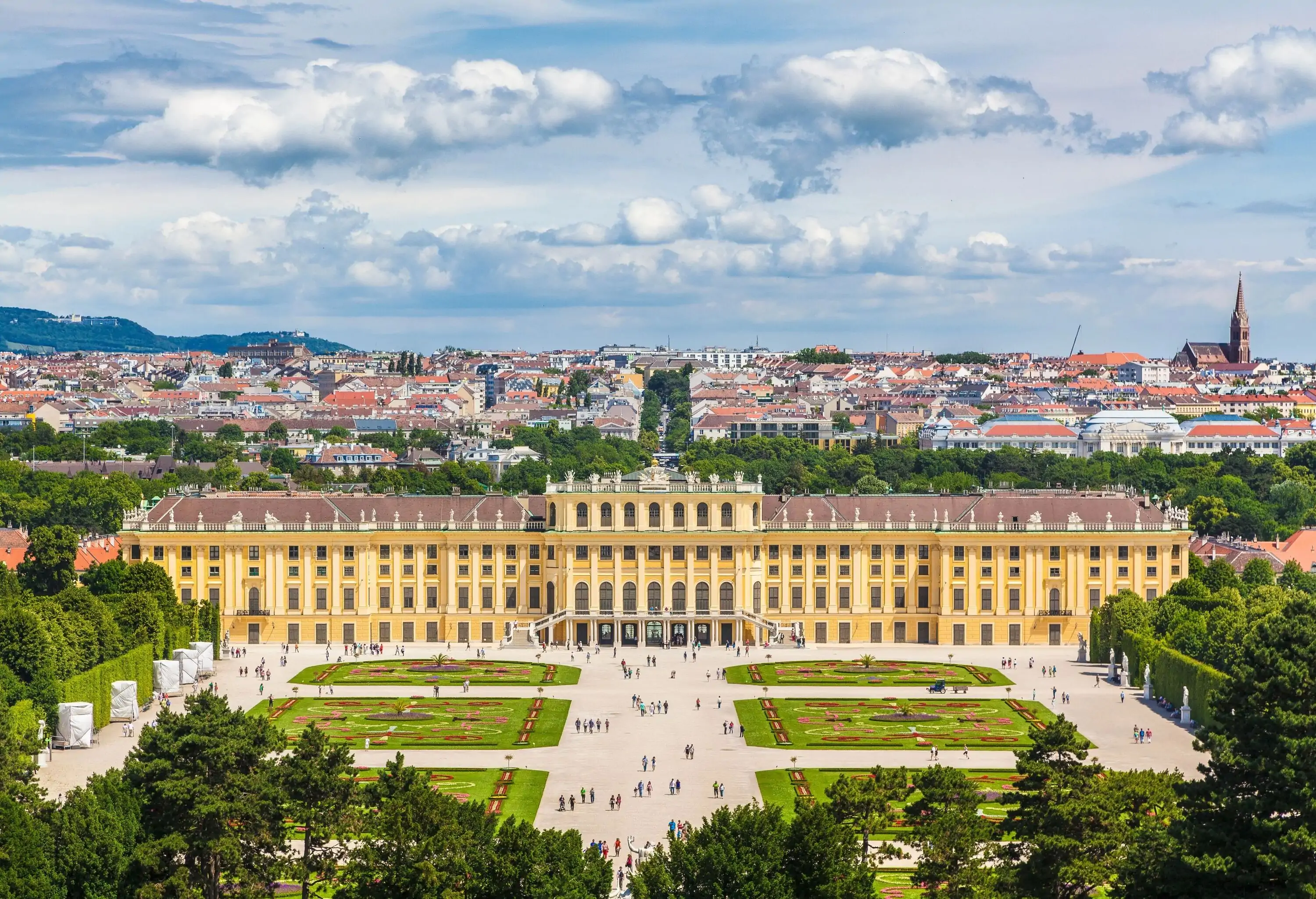 The grandiose Schönbrunn Palace, displaying ornate Baroque architecture with its imposing yellow facade, tall windows, and elaborate rooftop statues, stands majestically at the backdrop of its beautifully manicured Great Parterre garden, dotted with leisurely strolling visitors.