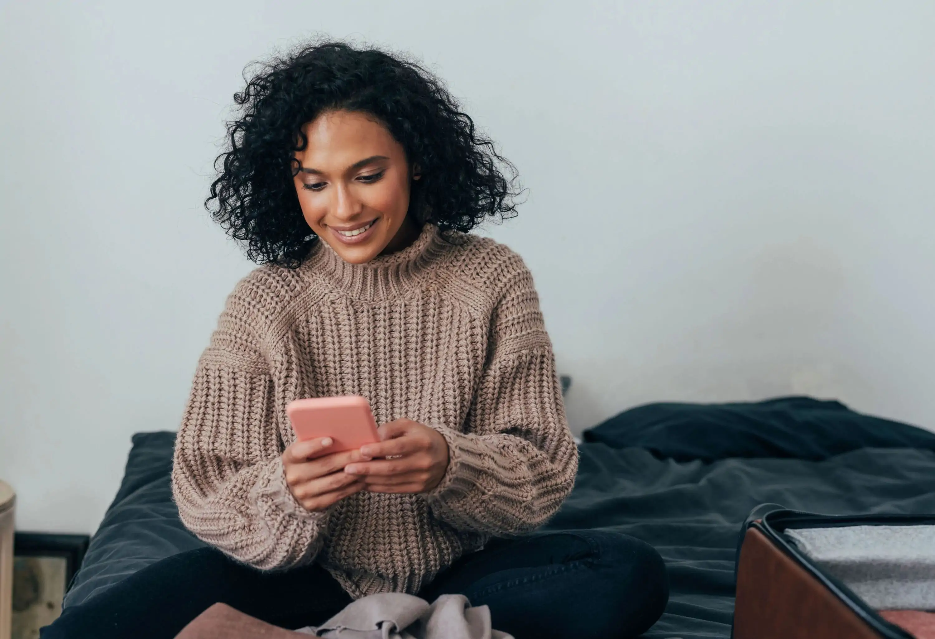 A woman with curly hair using her smartphone while sitting on her bed.