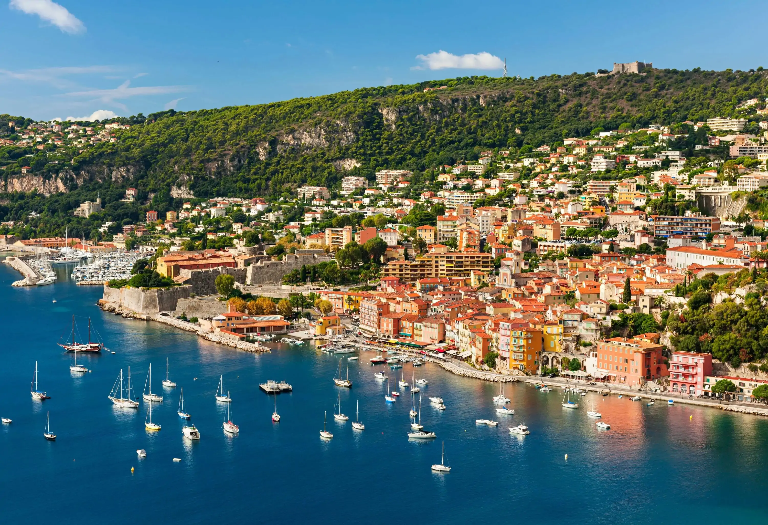 Sailboats dotting the surface of the azure sea, with buildings crowding the hillsides along the shore.