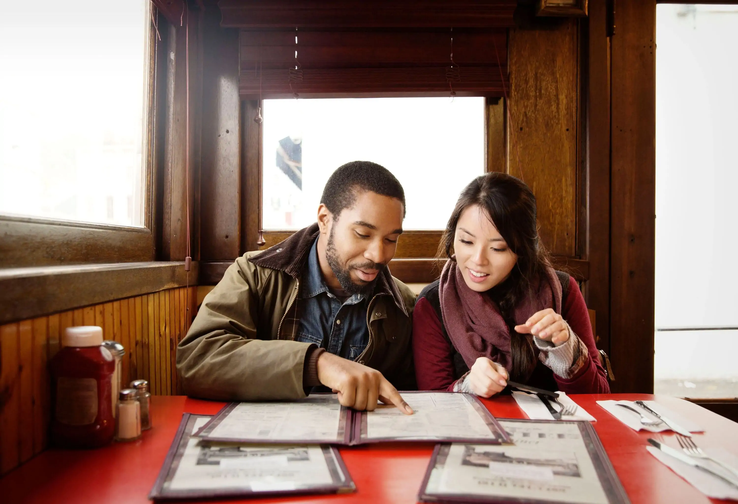 A couple wearing clothes suitable for cold weather peruses a menu at a diner with wooden walls, one pointing to an item while they make their selection.