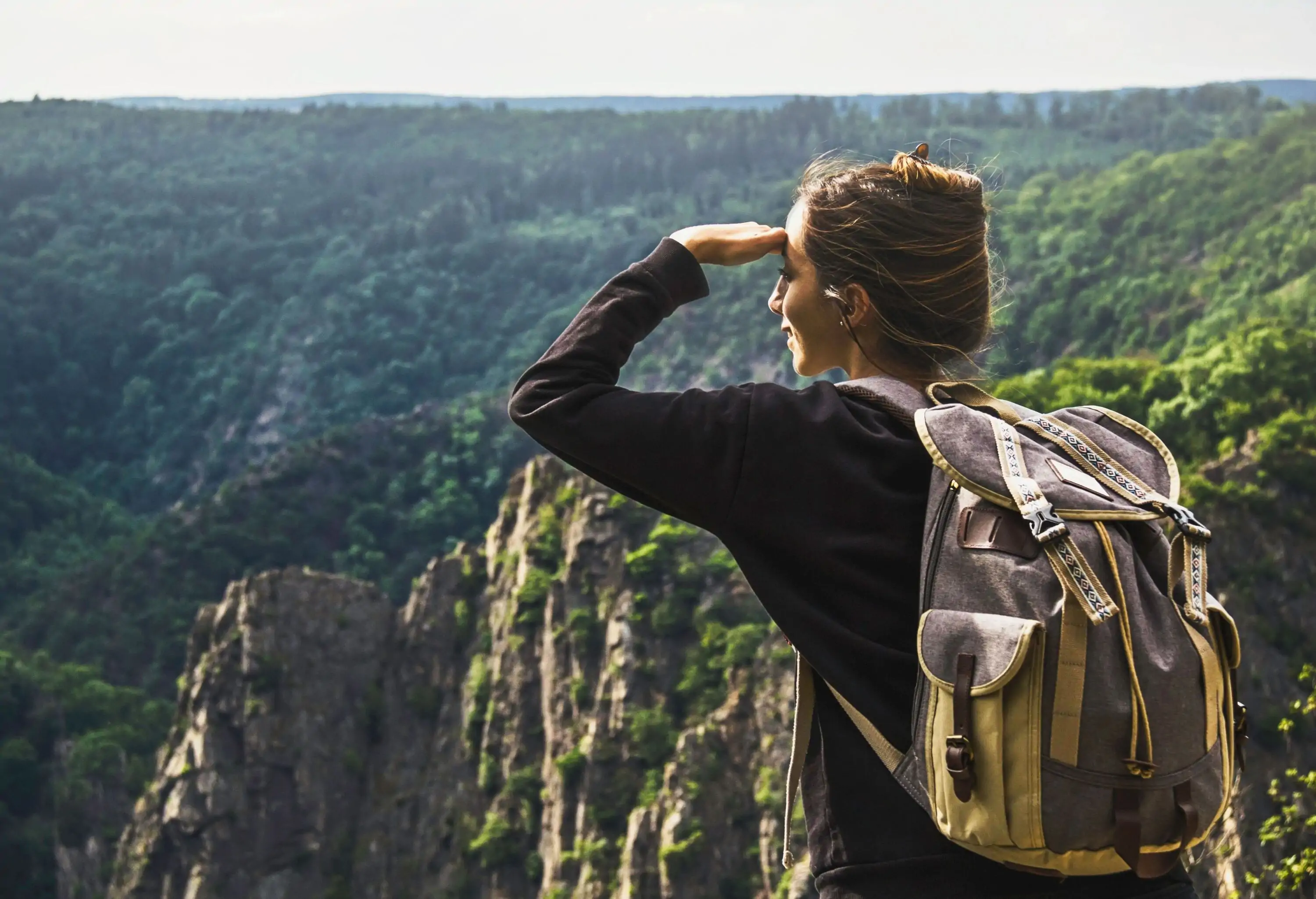 A woman in a black sweater and a big backpack stands on a summit overlooking the lush forested rocky mountains.