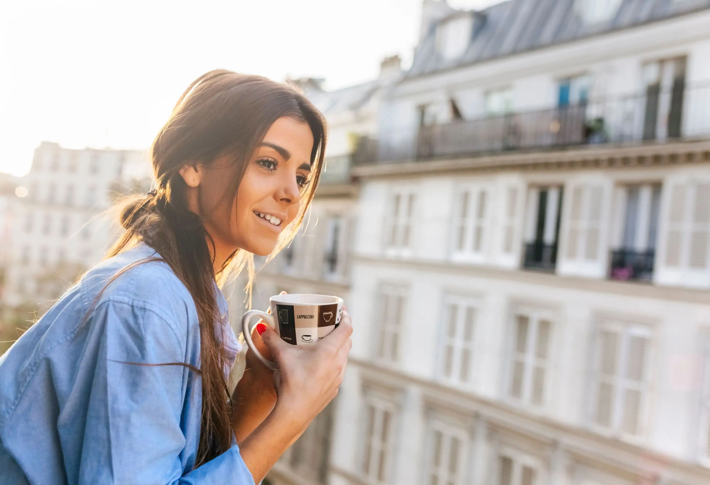 On the balcony, a woman with long black hair and blue long sleeves stands, smiling, and looks off into the distance while holding a coffee cup in both hands.