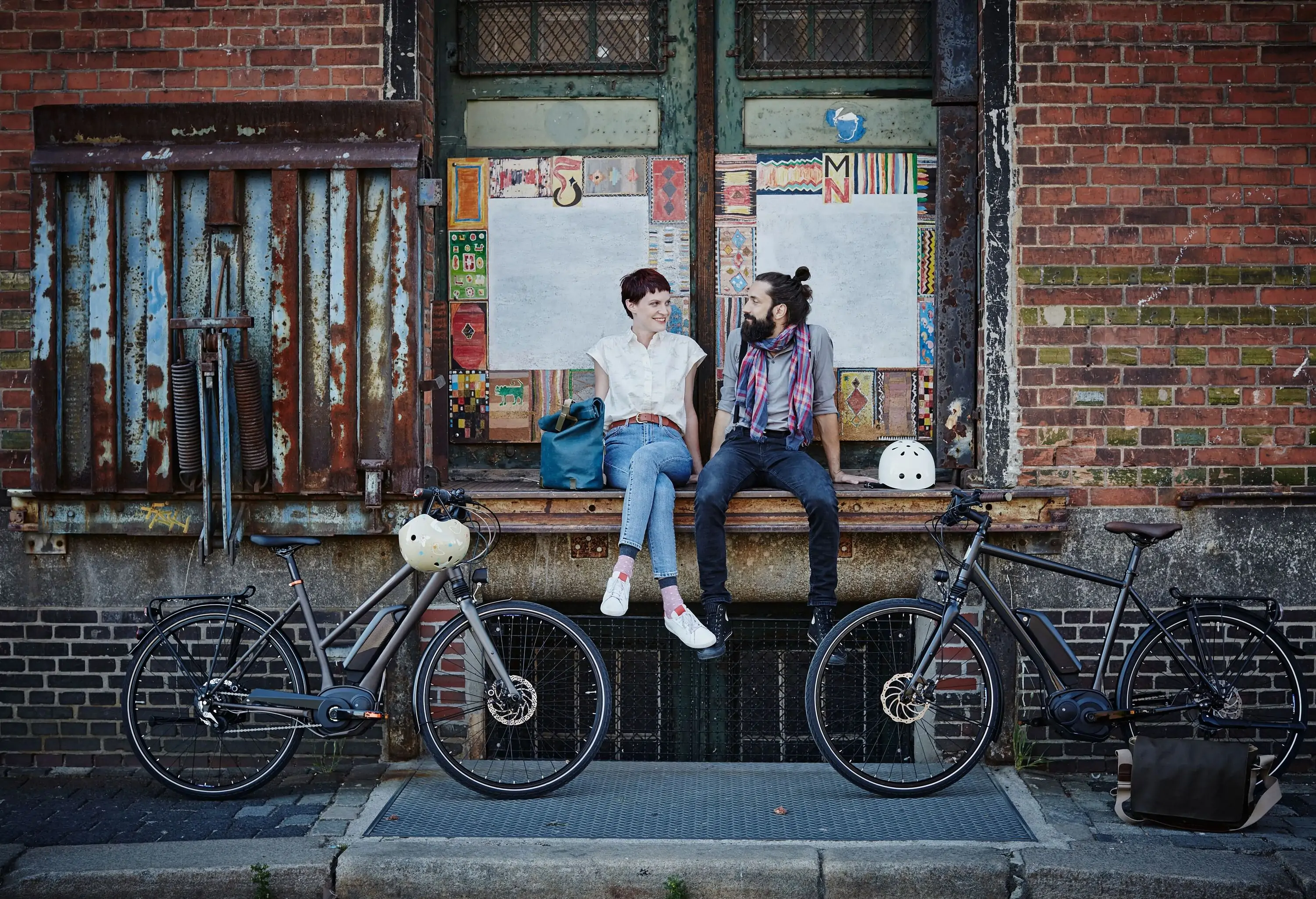 Two people in casual wear sit on a window ledge of a rustic and bricked building next to their bicycles.