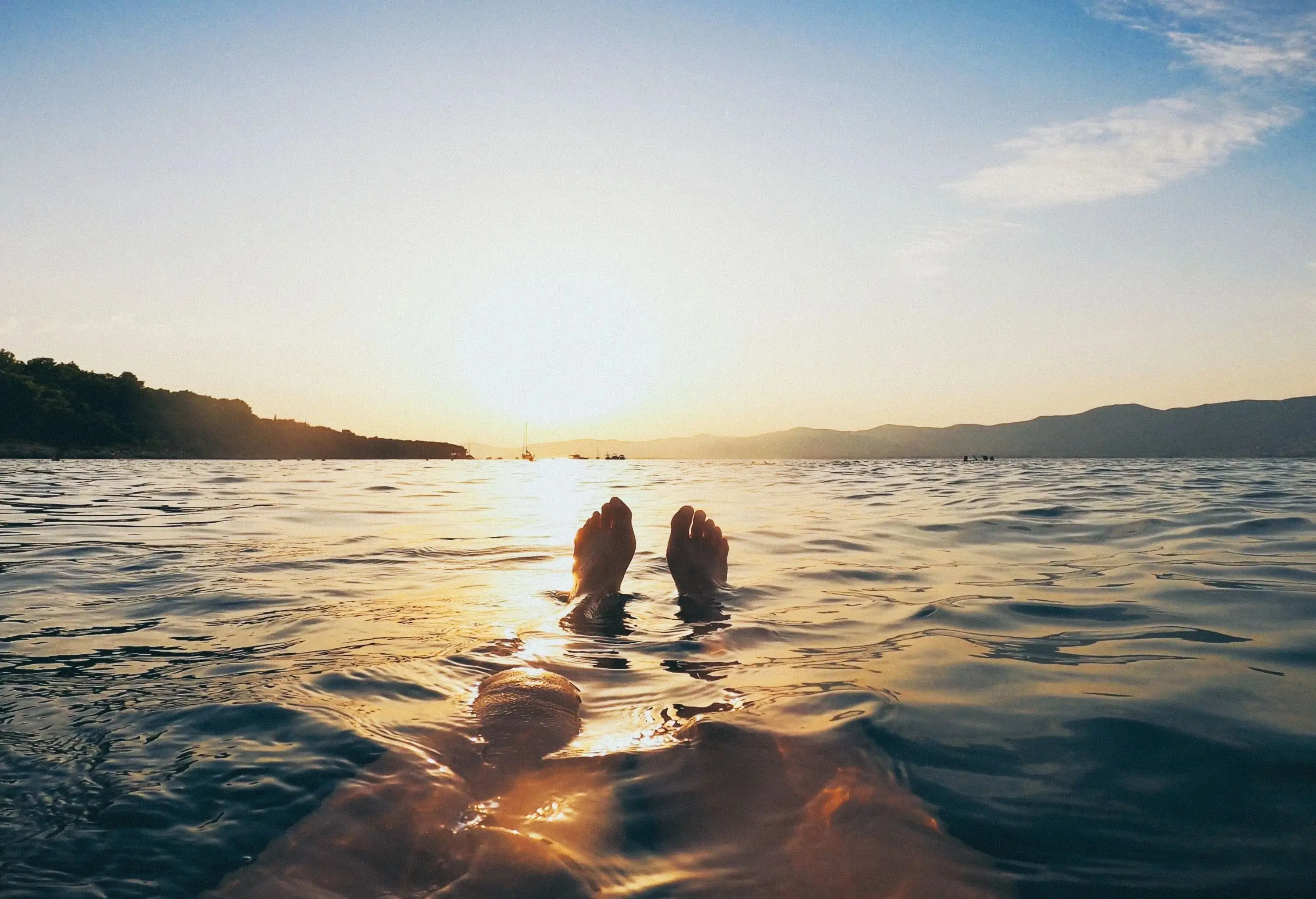 Sunlit bare feet submerged in the water with a view of steep mountain ranges against the bright blue sky.