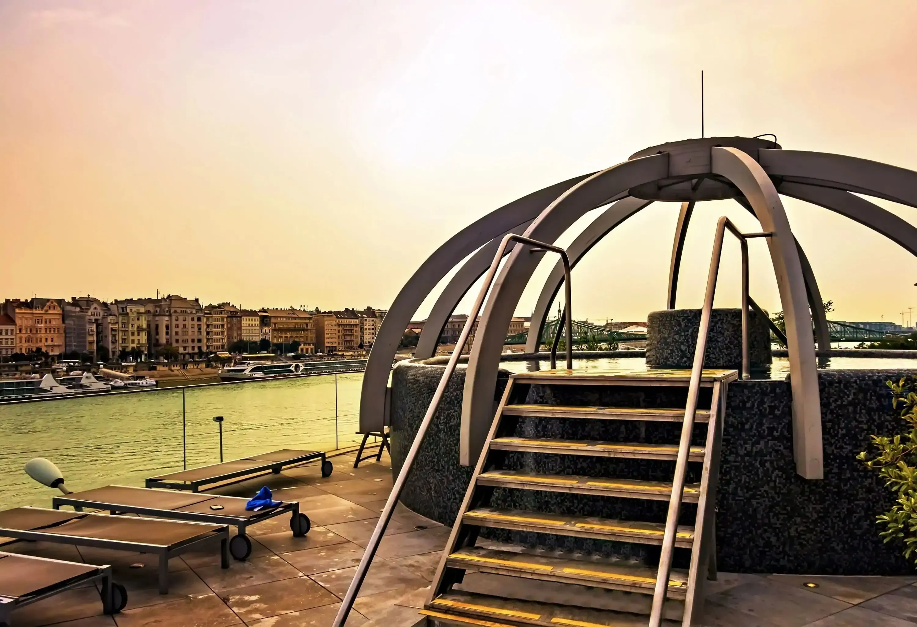 An octagonal rooftop thermal pool sheltered in a dome and accessed through a stair overlooking the river and tall buildings.