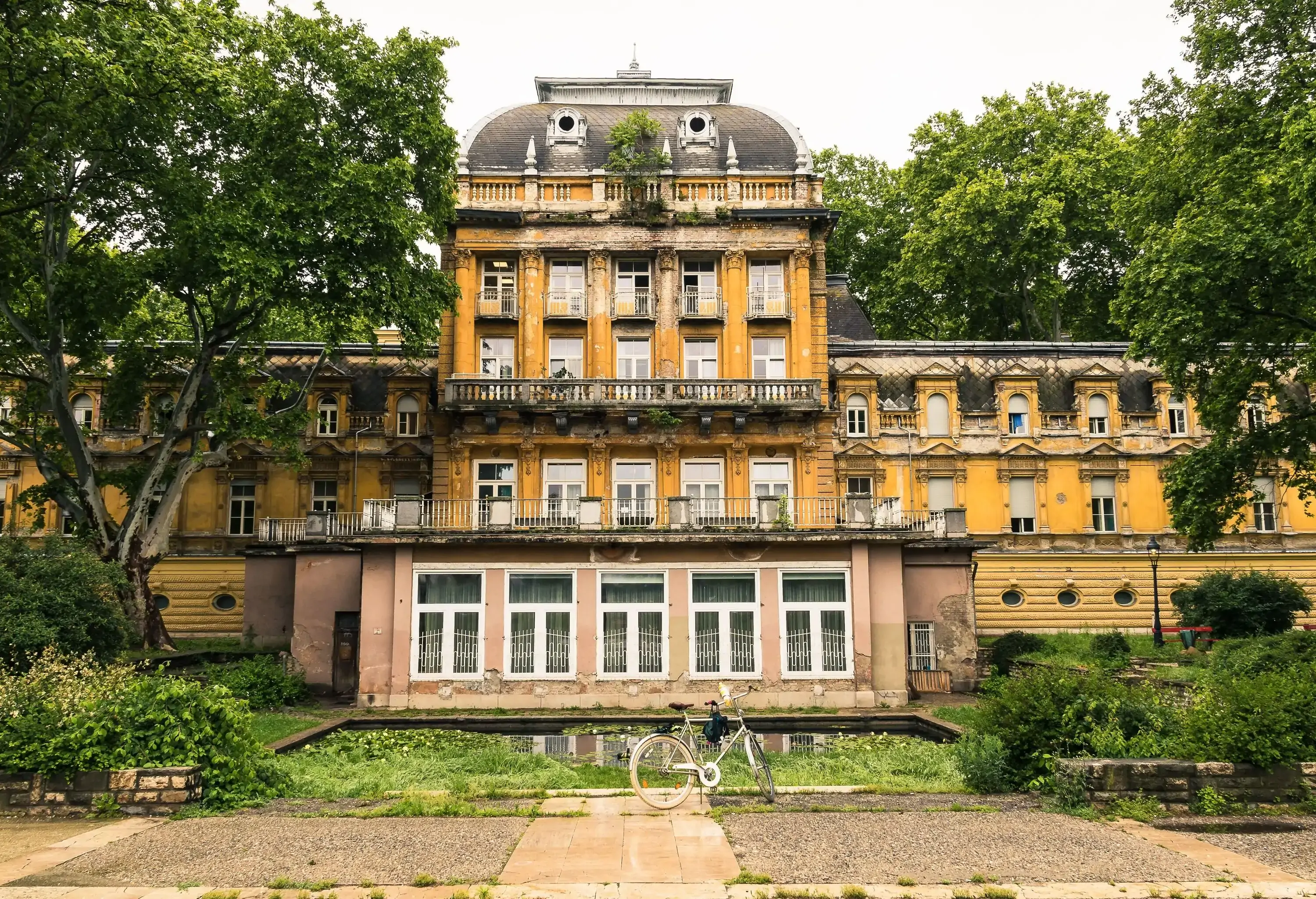 A bicycle parked in front of Lukács Thermal Bath, a thermal bath spa with a neoclassical façade of multiple windows and pillared balconies.
