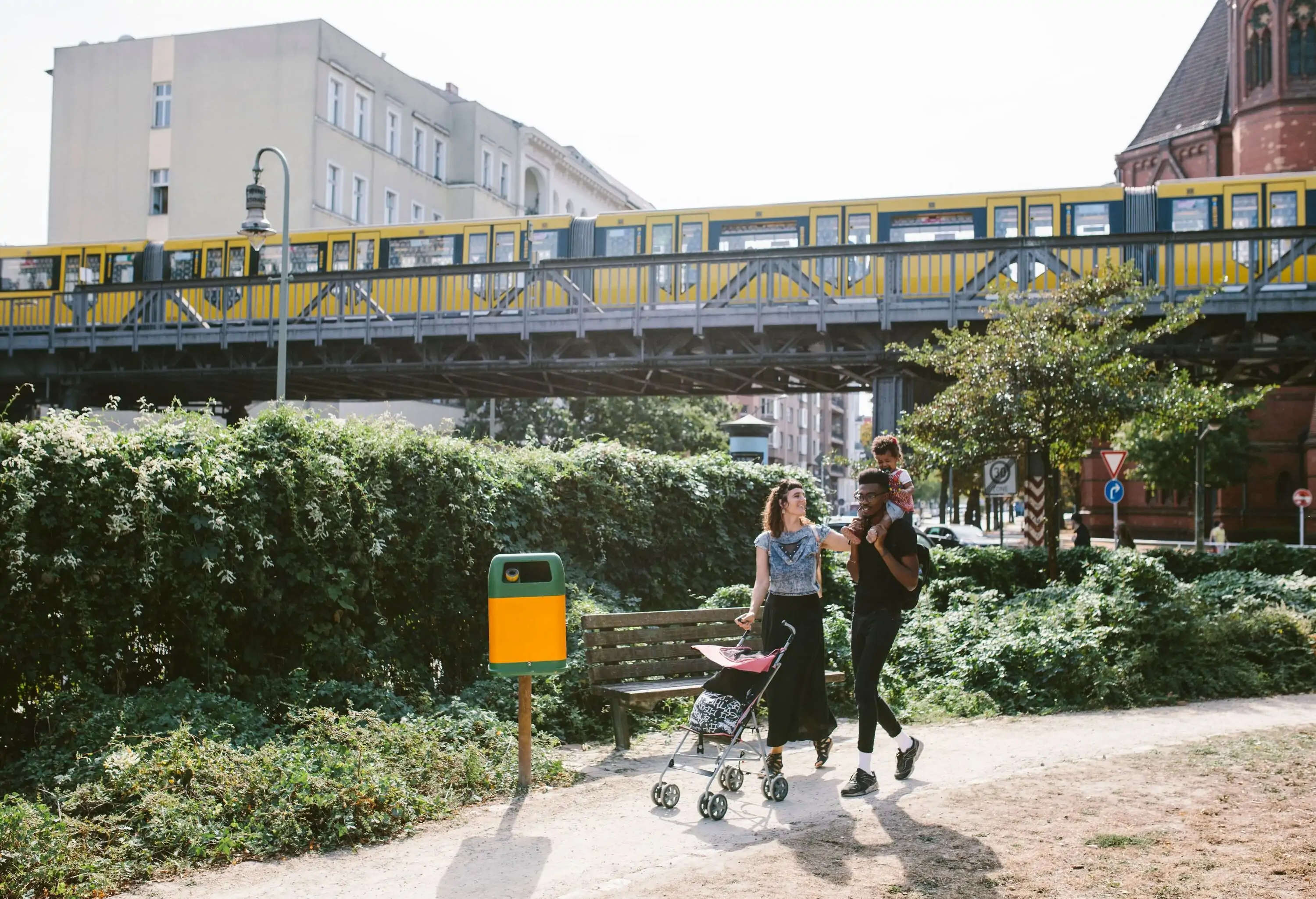 A father carries his toddler on his shoulder alongside her wife, walking on an unpaved pathway with a yellow train travelling above them in the background.