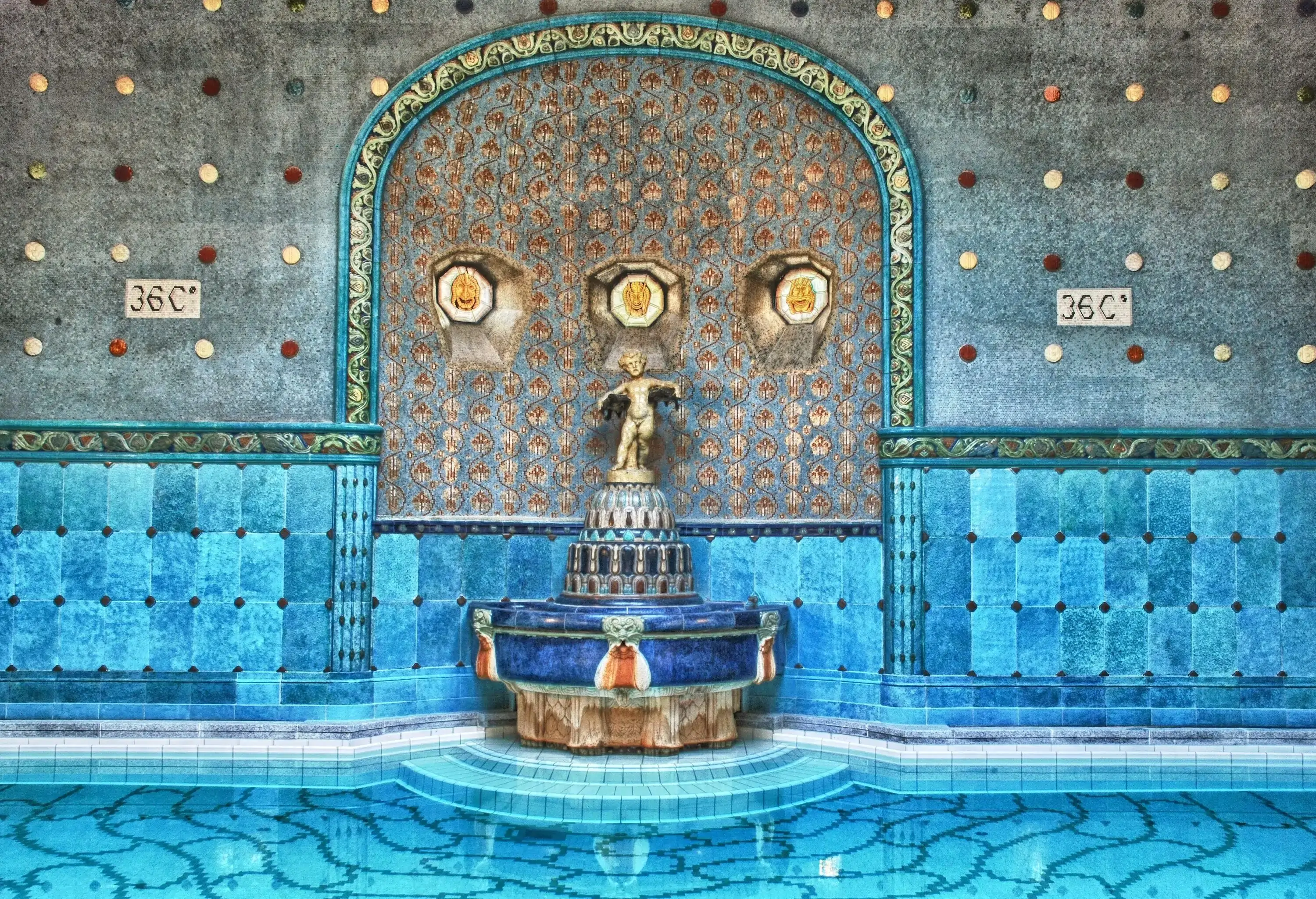 An arch wall with turquoise mosaic tiles, ornately-carved sculptures, and ceramics dotted everywhere in a thermal bath.