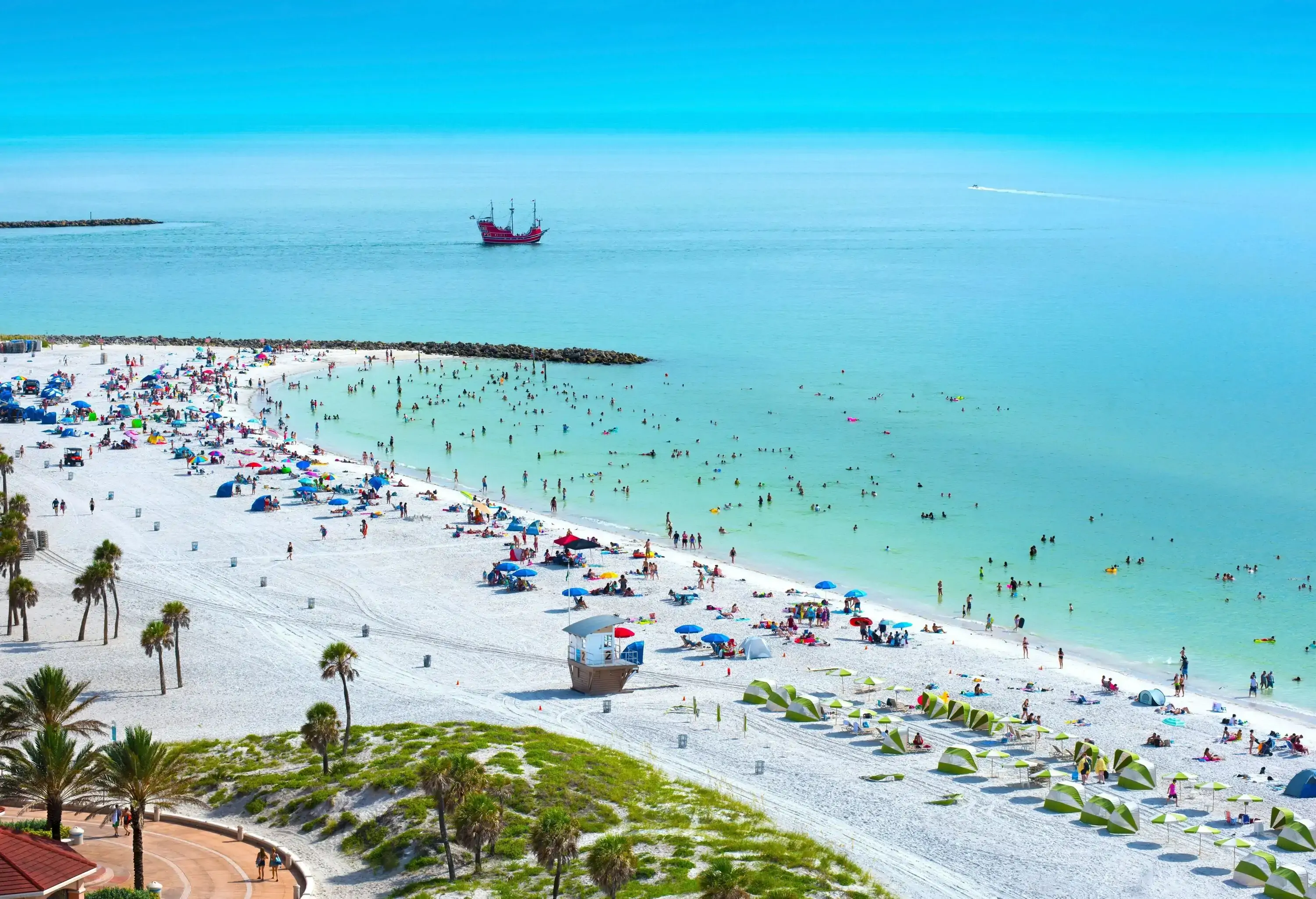 A bustling Clearwater Beach teems with visitors as colourful parasols and sunbeds dot the shoreline, while the sea shimmers in the sun and a sailing ship sails majestically in the distance.