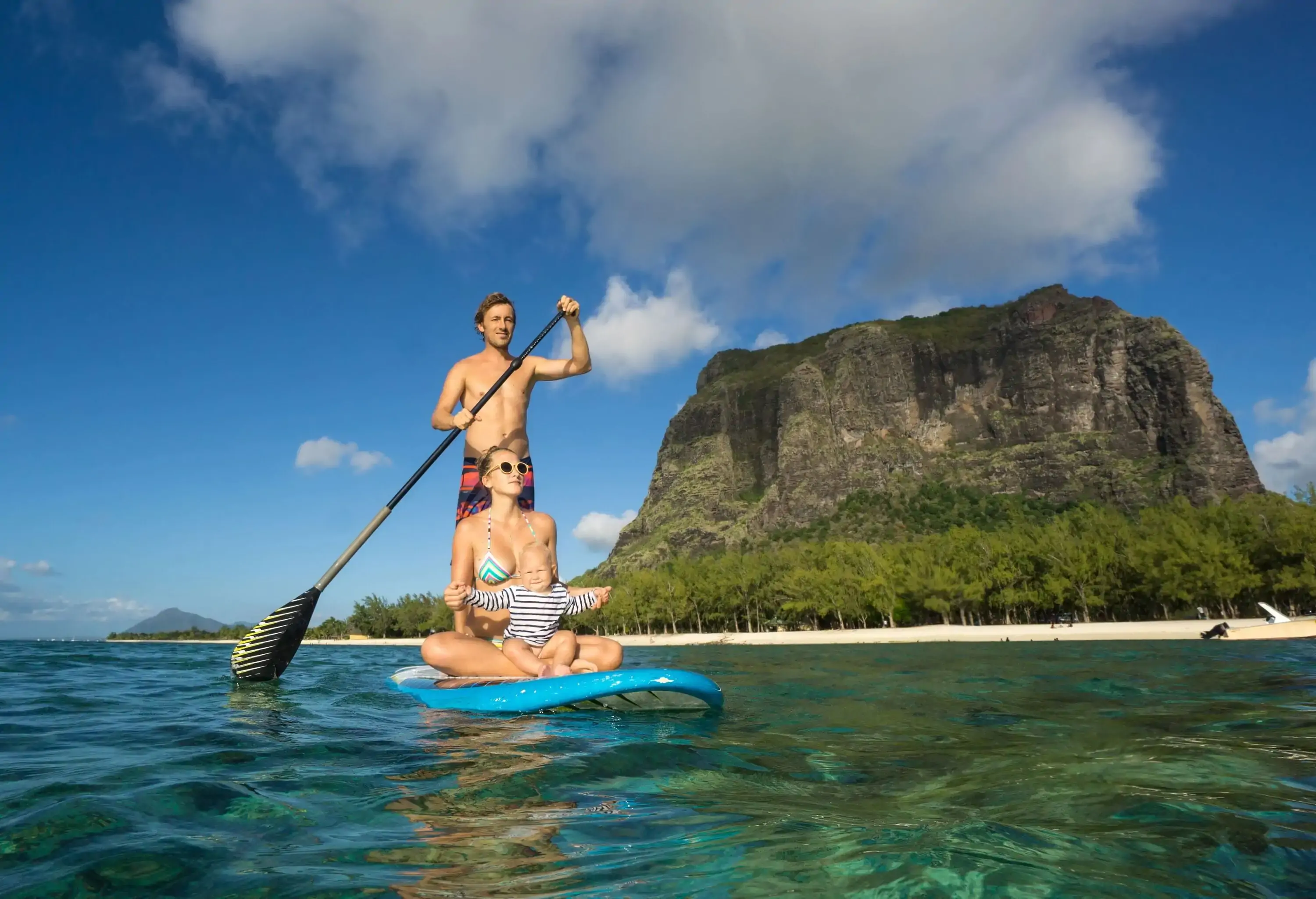 A man stands on a surfboard paddling beside a sitting mother and infant, with views of a mountain in the distance.