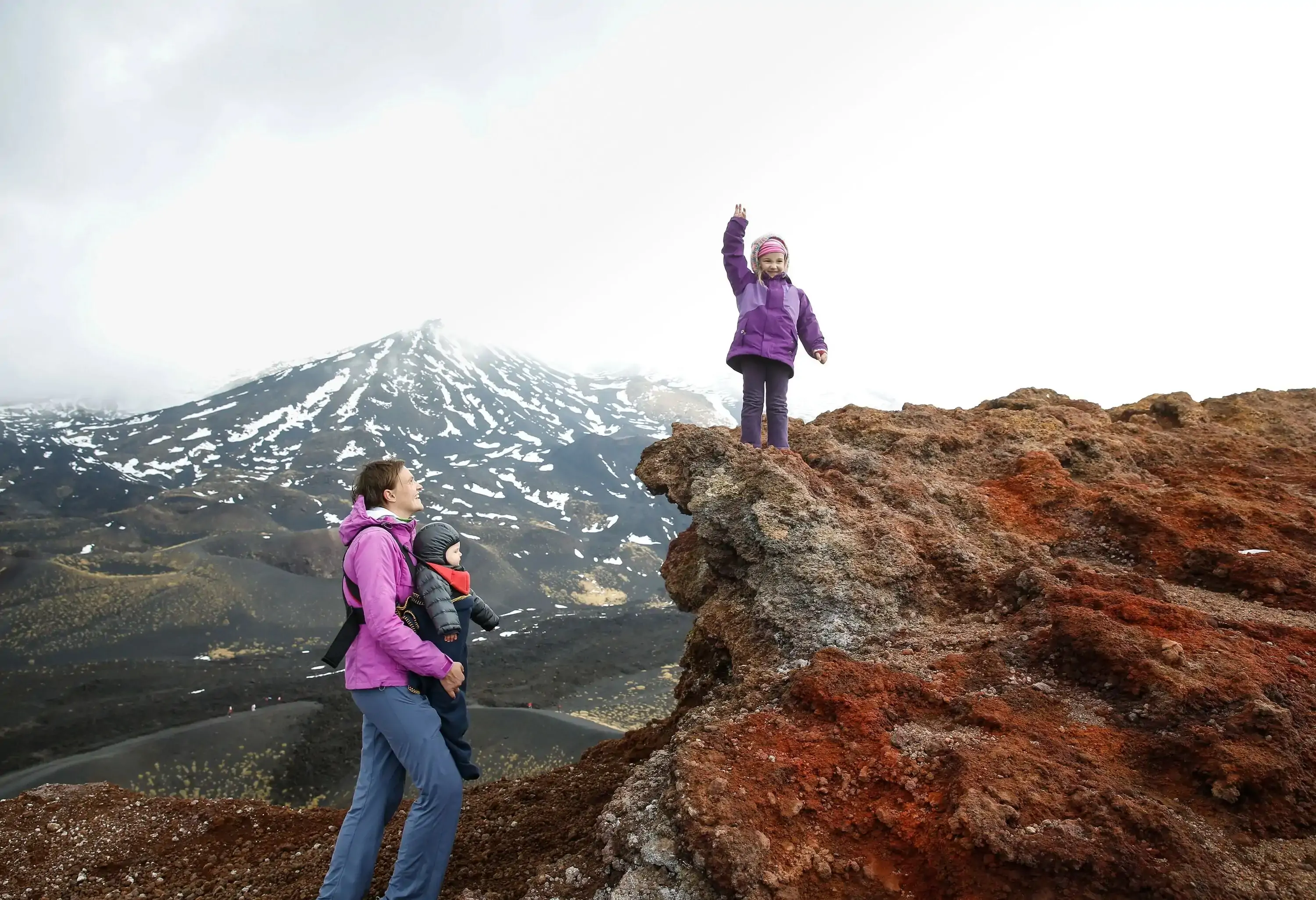 Mother with her baby and triumphant toddler on the edge of a crater on Mount Etna, volcano on the island of Sicily, Italy. Active family lifestyle, traveling family concept.