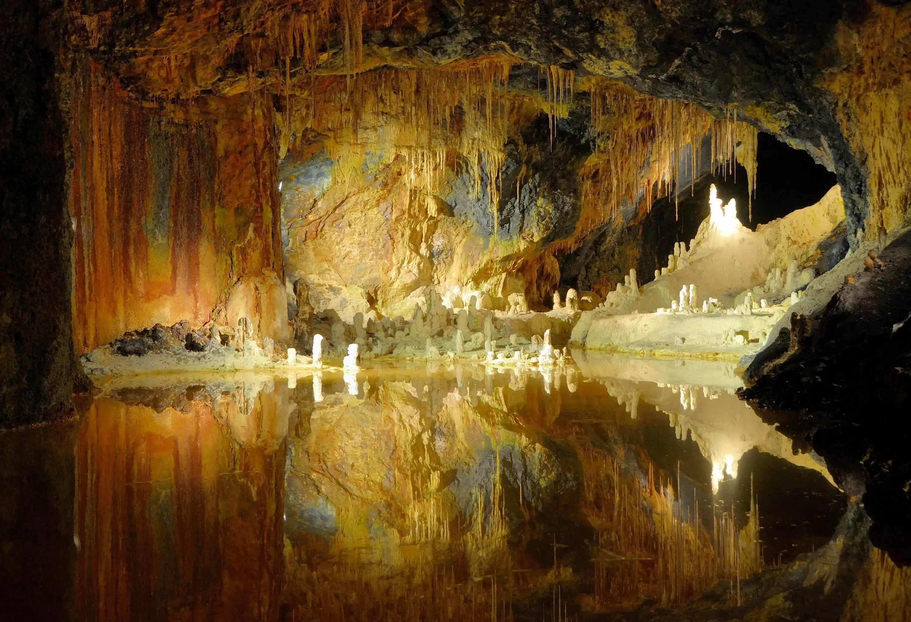 A cave with a body of water reflects the brightly-lit stalagmites and stalactites.