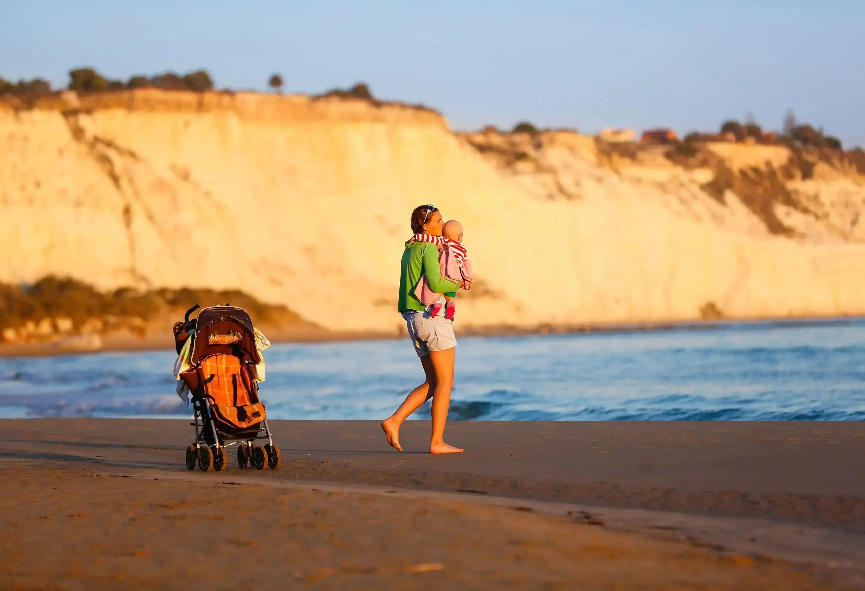 Affectionate mother holding and kissing her baby boy on a sandy beach, enjoying the cool of late summer evening. Family vacation, traveling with children concept.