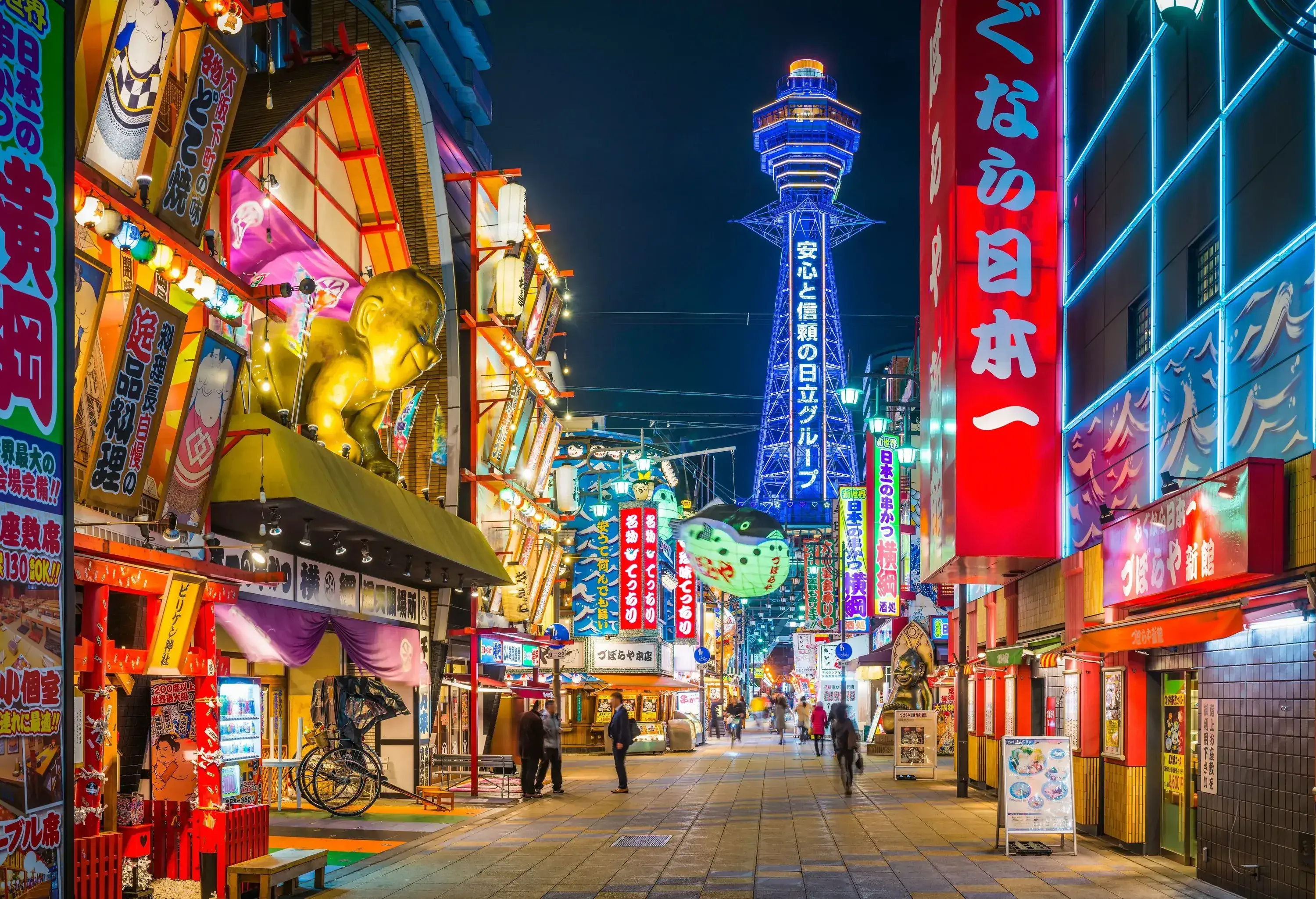 A street in a neighbourhood across commercial establishments illuminated with neon lights and views of an observation tower in the distance.