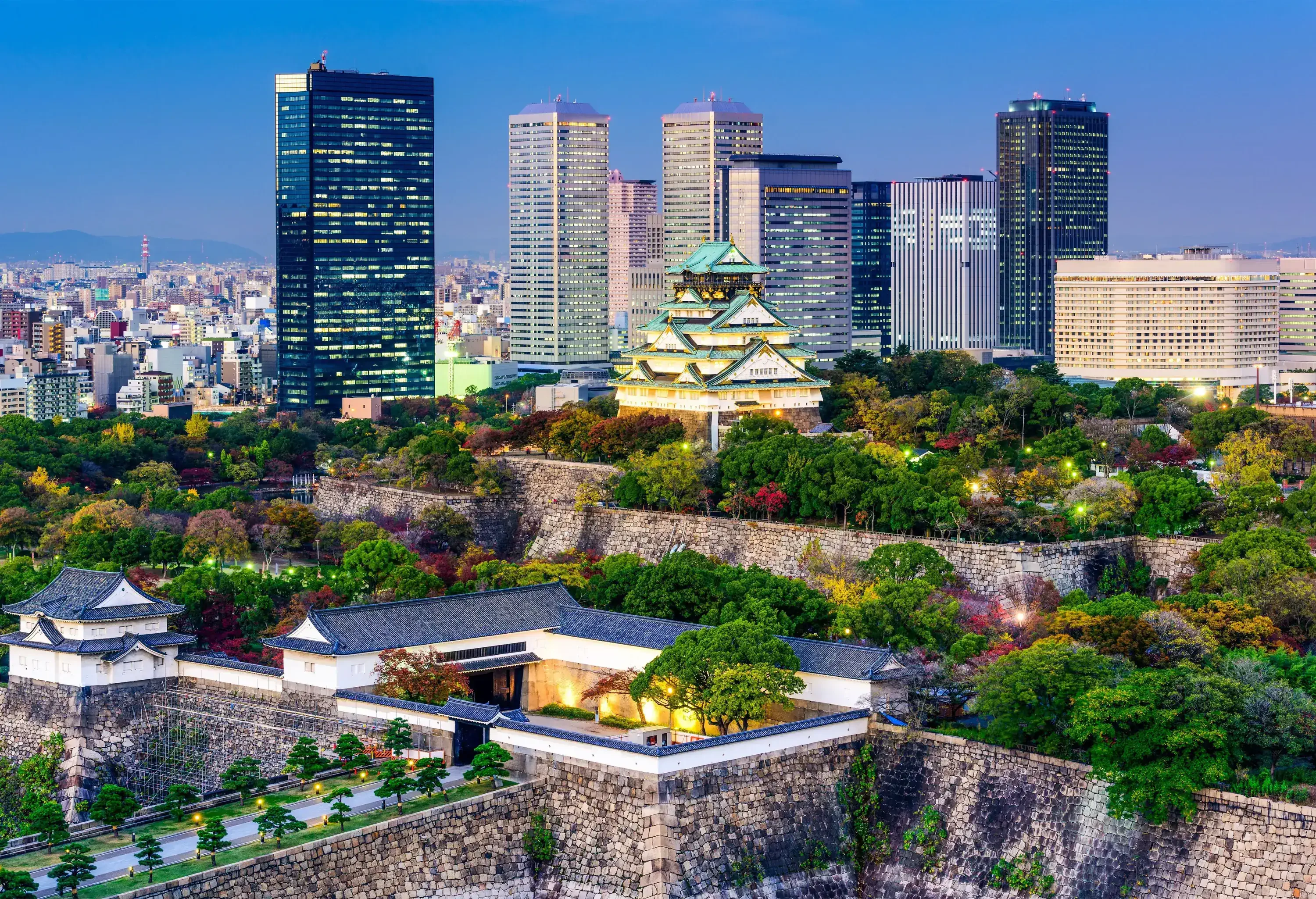 Multicoloured autumn trees surround the illuminated Osaka Castle, with a modern skyline and metropolis in the distance.