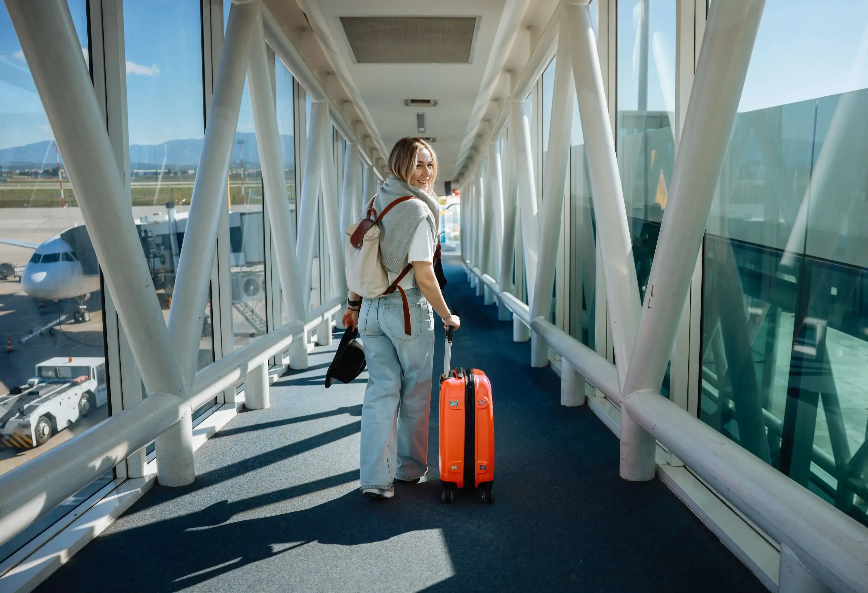 Close-up image of attractive, female traveller wearing casual clothing, cropped top, pushing wheelie suitcase luggage with telescopic handle over glass walled jetway loading bridge to board airplane, focus on foreground