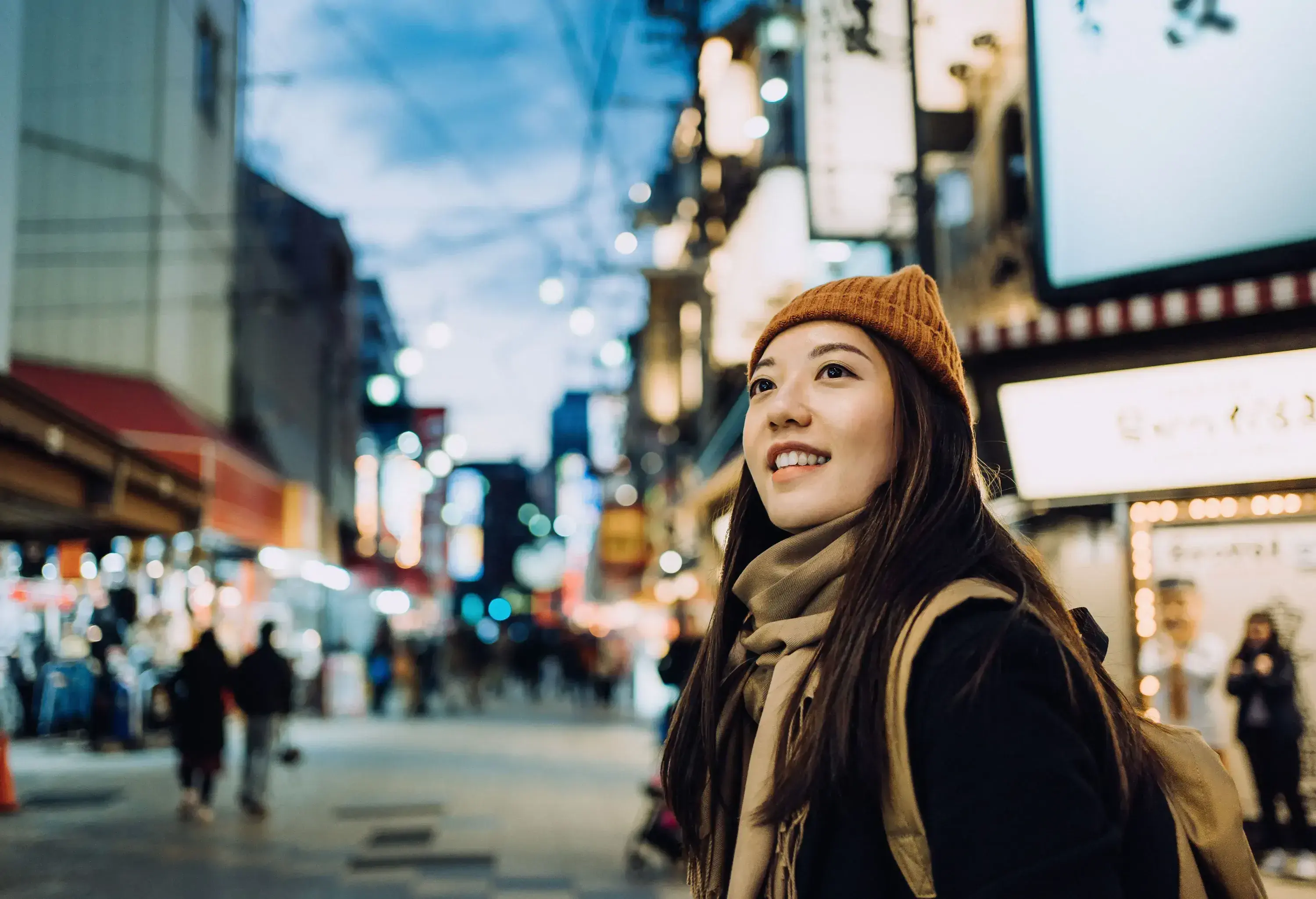 A smiling young Asian female traveller, sporting a beanie, scarf, and black coat, delightfully explores and strolls through a bustling local city street, soaking in the vibrant atmosphere of her surroundings.