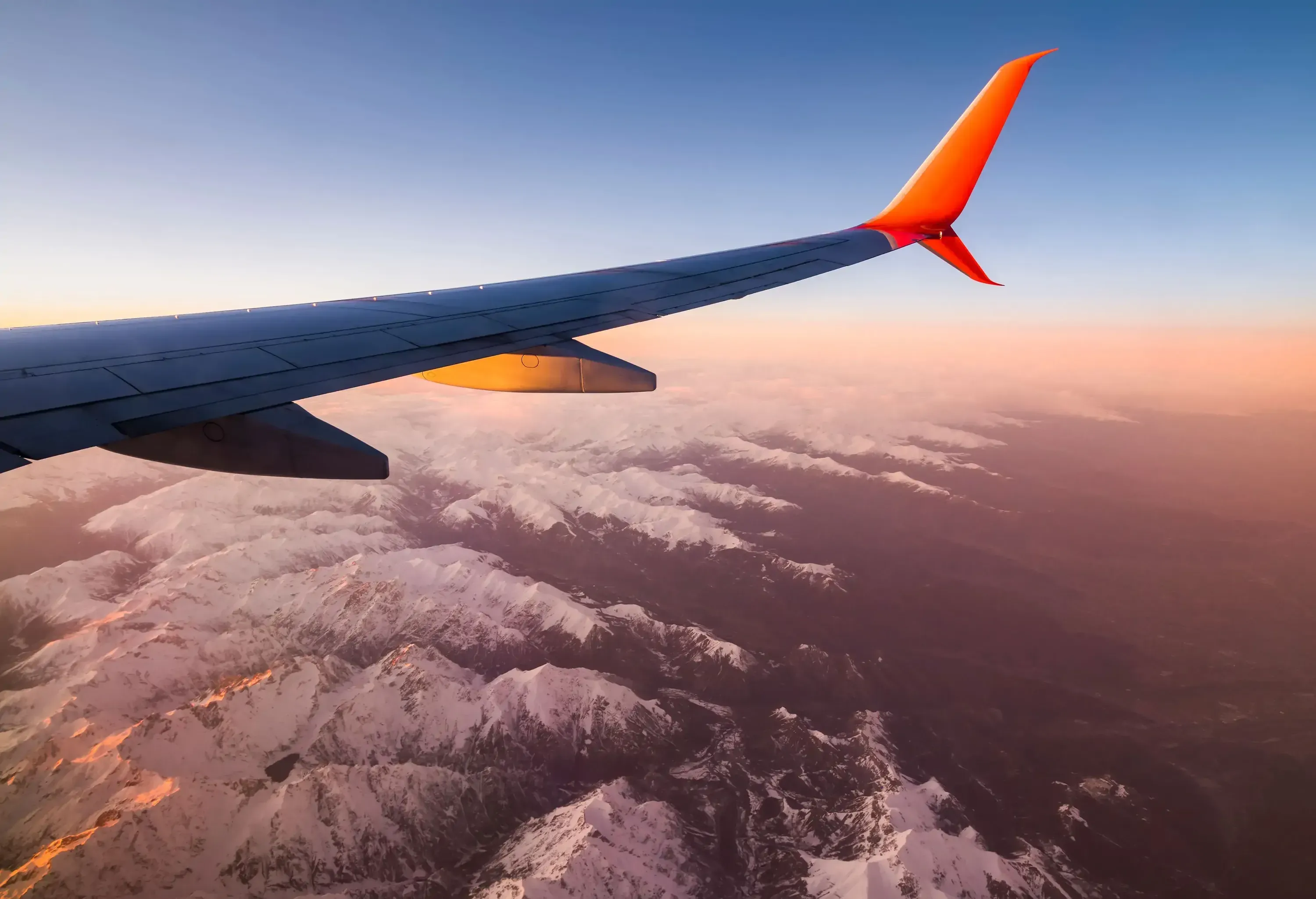An airplane wing against a sunset sky, with snow-covered mountains below.