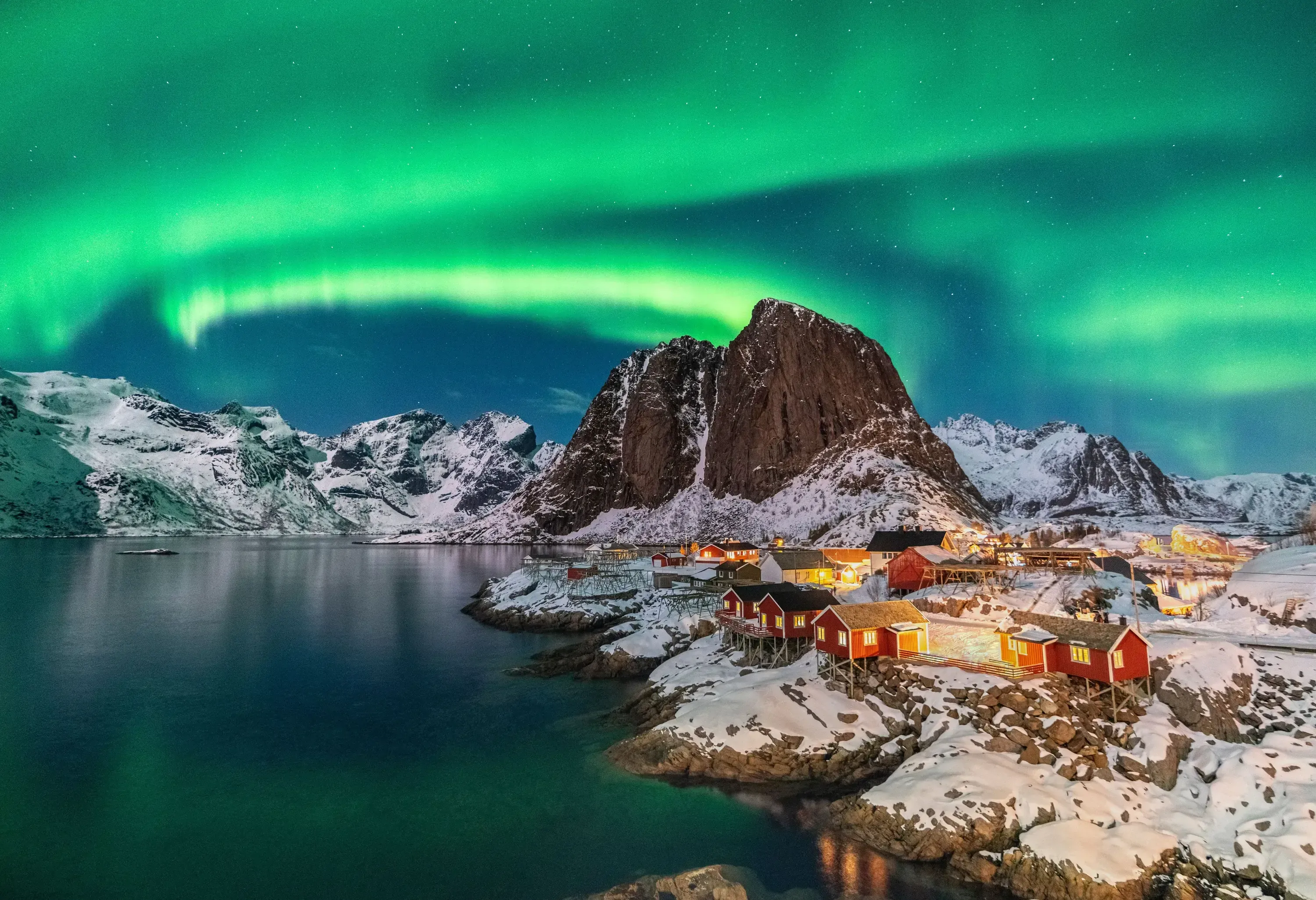 The dazzling Northern lights colour the sky green and blue over a small village along the lake bounded by steep rocks.