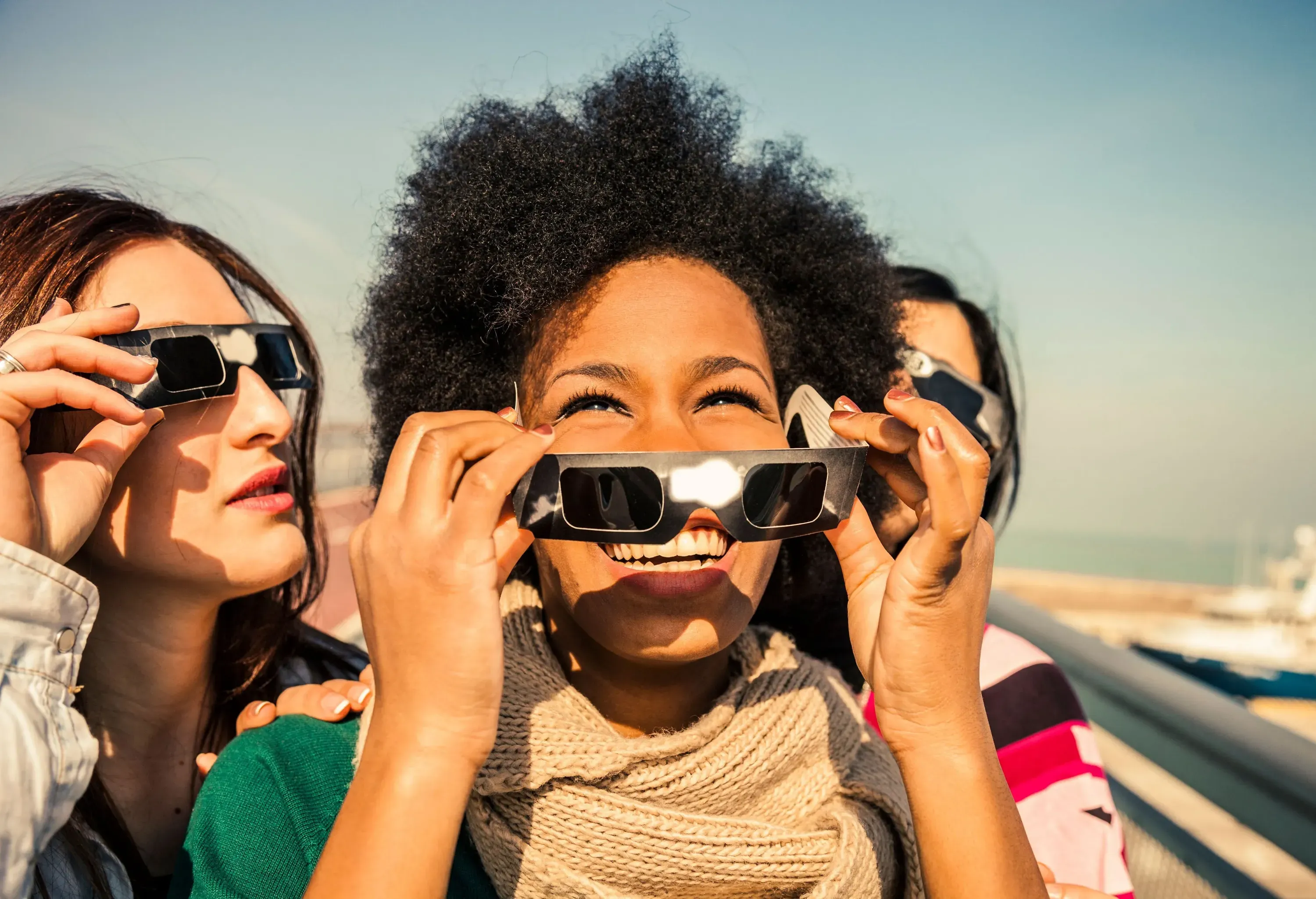 Three people gaze upwards through eclipse glasses under a bright sky.