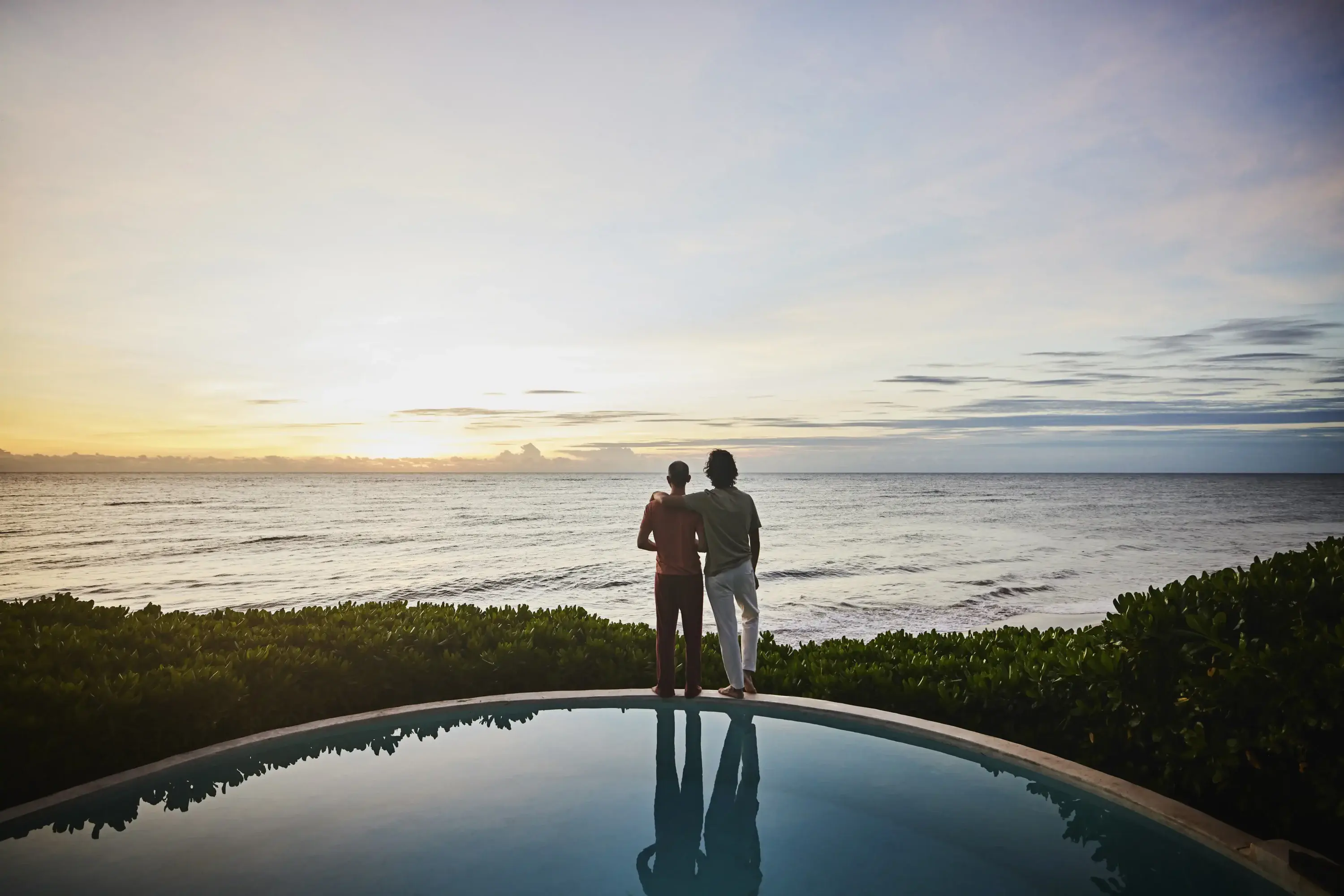 A couple stands on the edge of a pool, overlooking the distant sea, with lush green surroundings visible in the background.